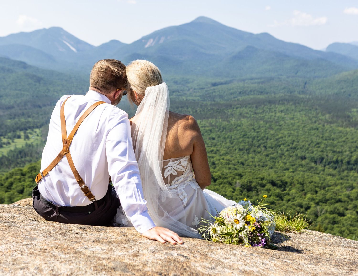 Newlywed couple sitting on a mountain rock overlooking lush green valleys, bride holding wildflower bouquet in wedding attire.