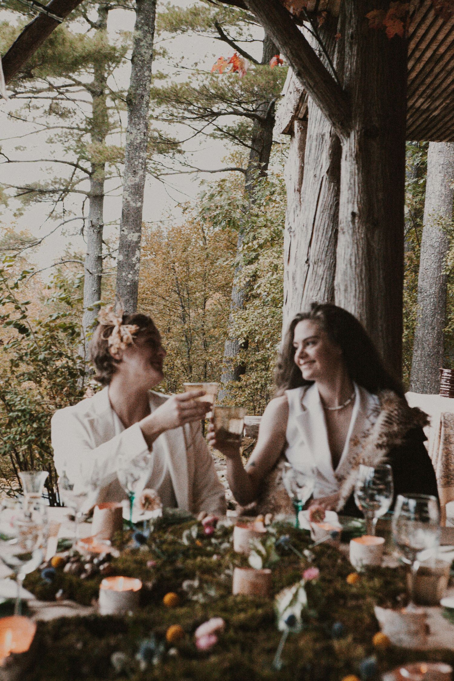 Wedding reception guests enjoy a meal at long tables decorated with flowers under tall pine trees.