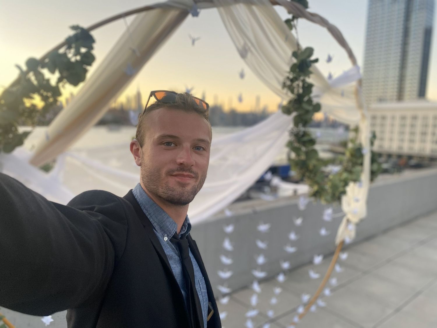 Young man taking a selfie in front of a decorated wedding arch on a rooftop at sunset.