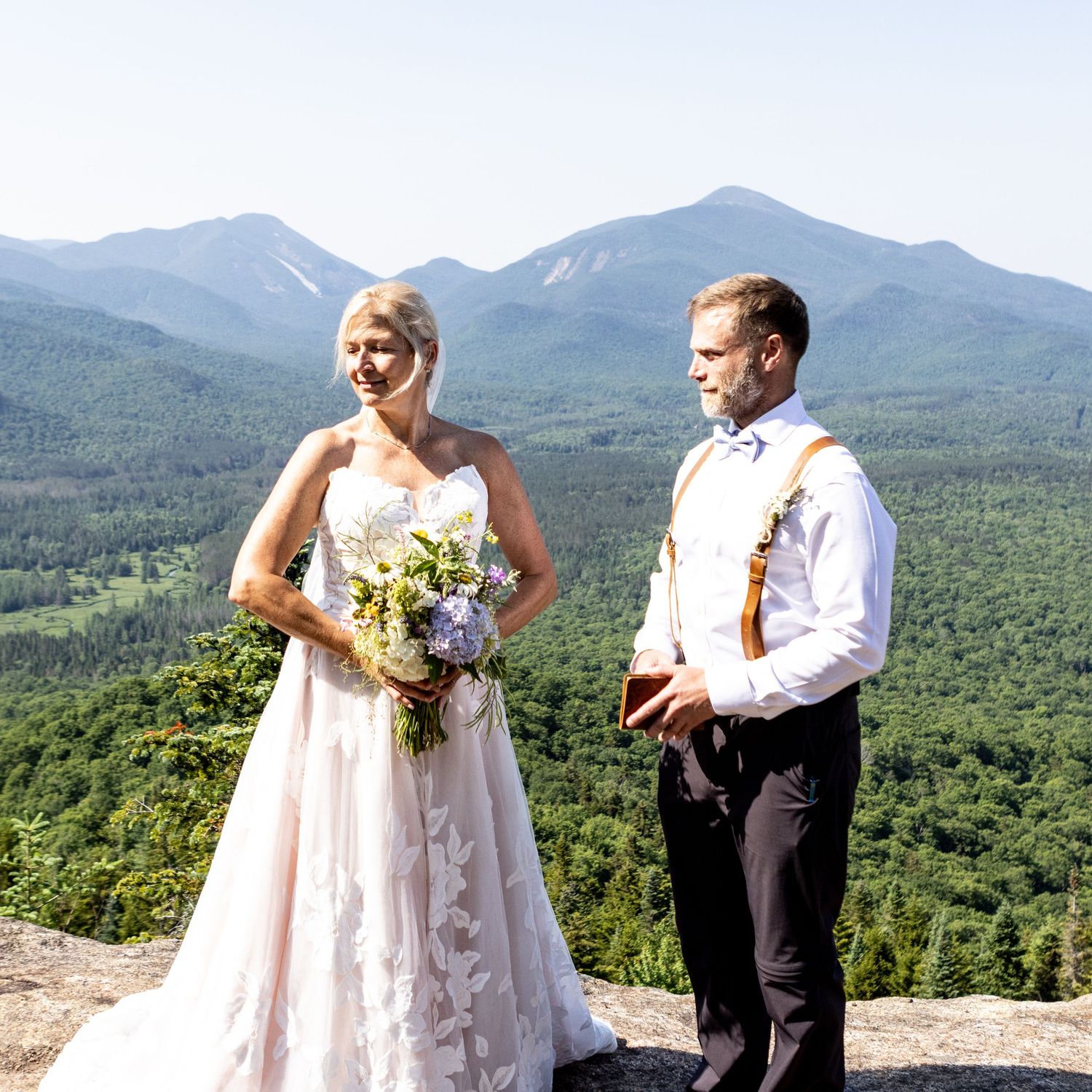 Bride in white lace gown holding bouquet stands with groom in suspenders on mountain overlook with scenic valley views.