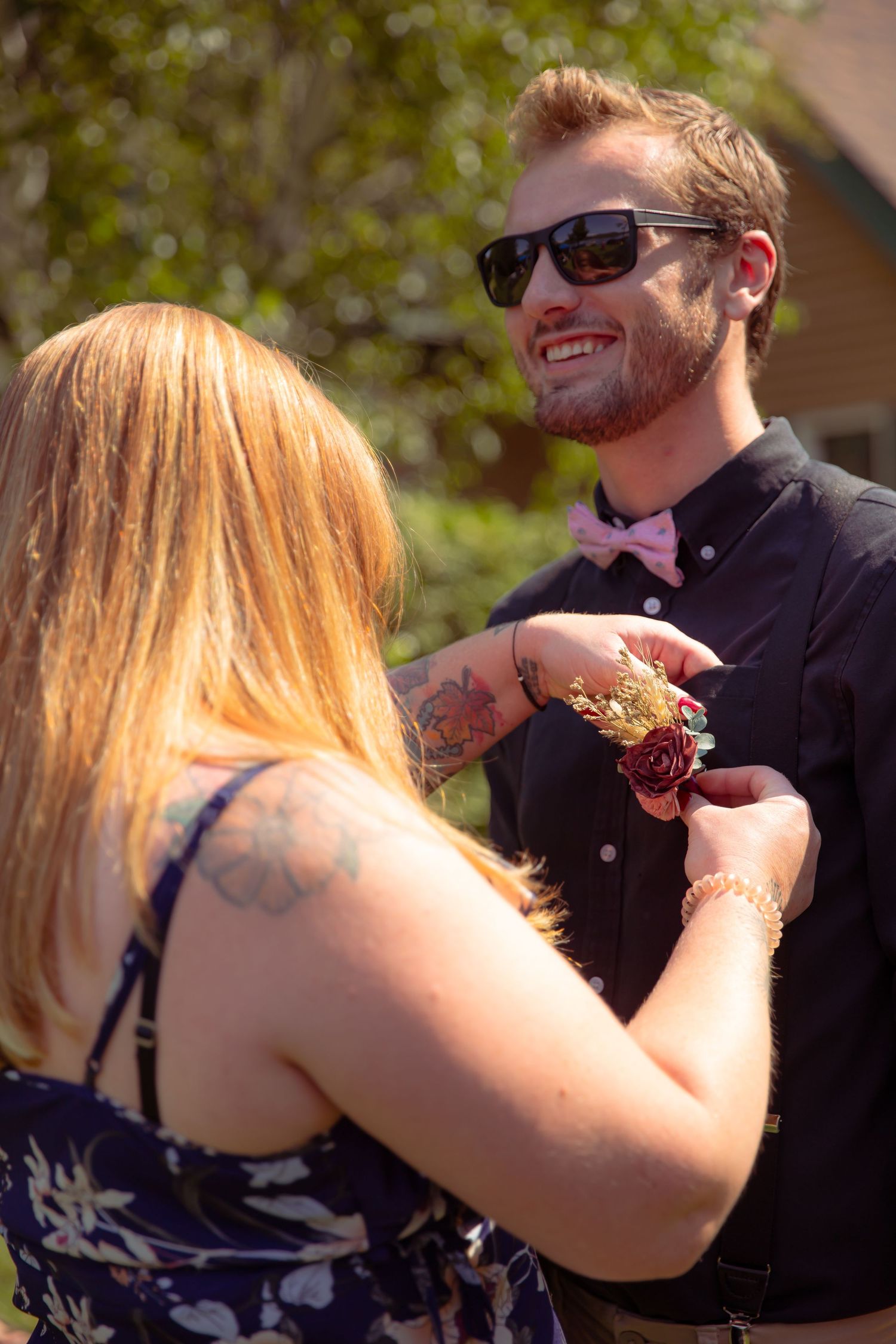 Young woman with tattoos pinning a boutonniere on a smiling man in sunglasses and pink bow tie outdoors.