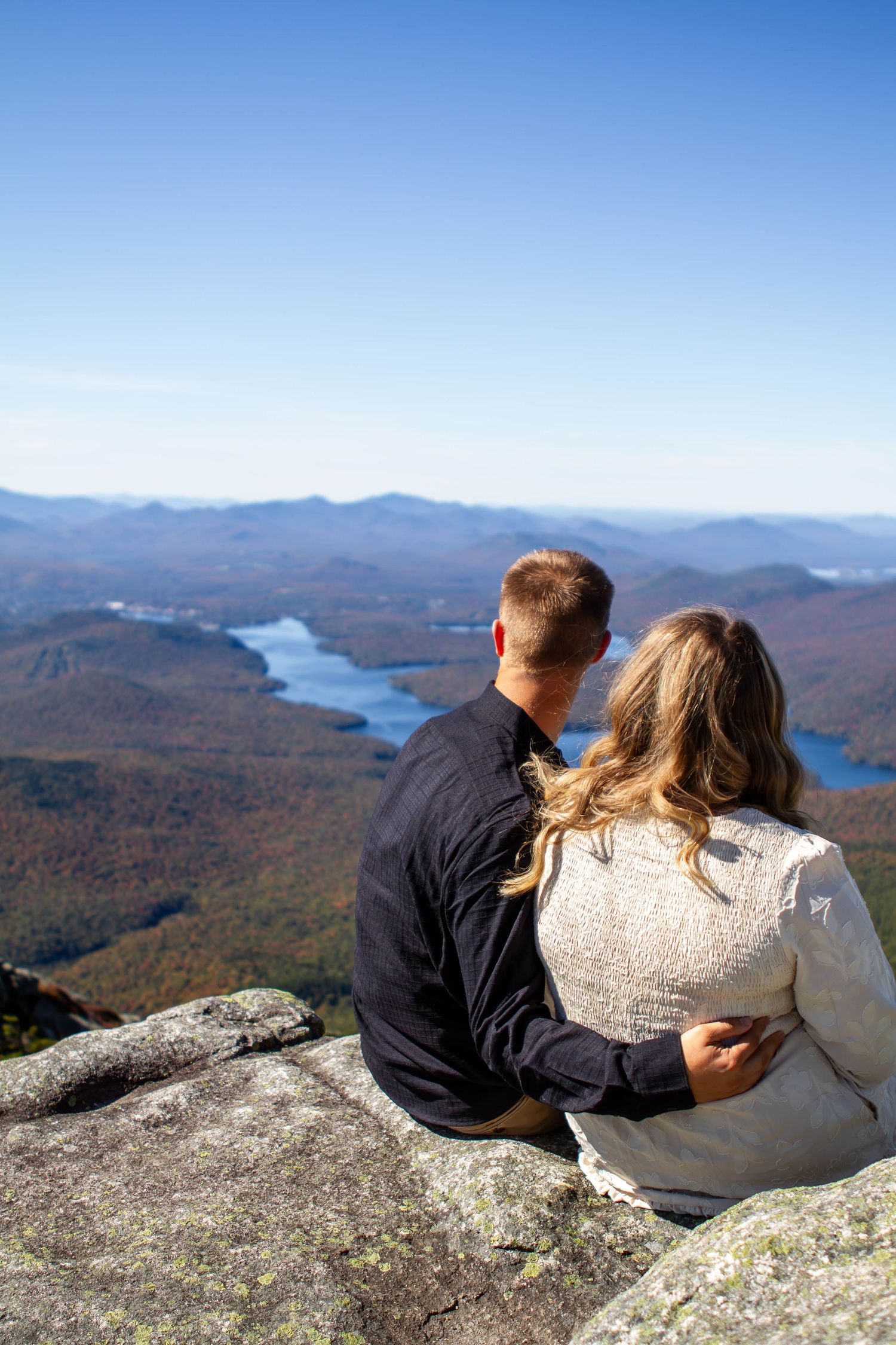 A couple sits on a rocky mountain overlook enjoying scenic views of a lake and autumn forest landscape.