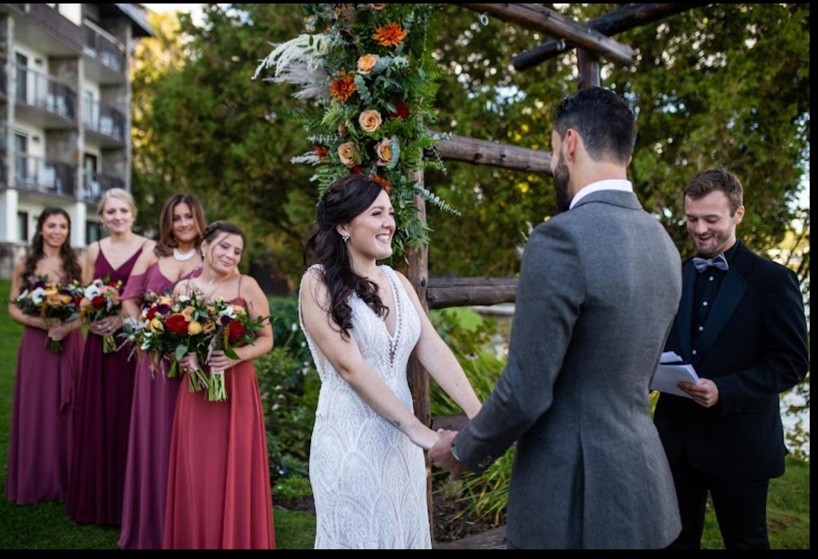 A wedding ceremony takes place outdoors with attendants in burgundy dresses and floral decorations.