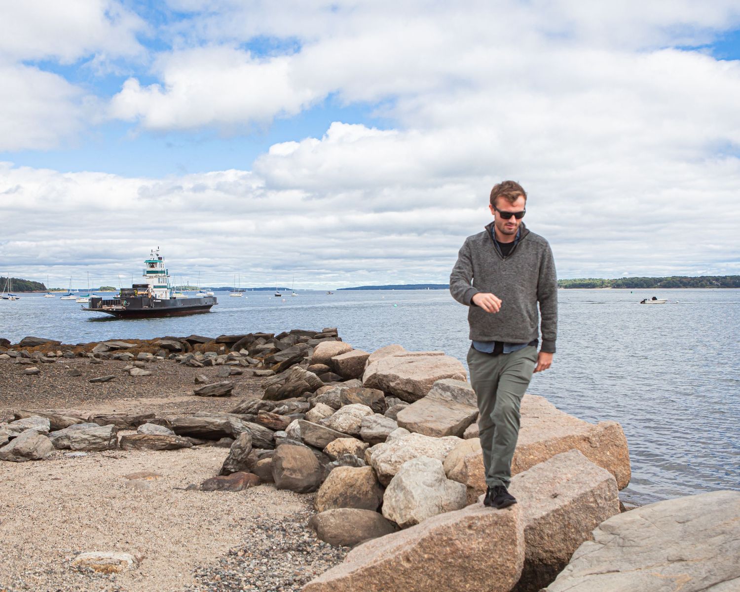 Man in gray sweater walking on rocky shoreline with a ferry boat and calm blue ocean waters in the background.