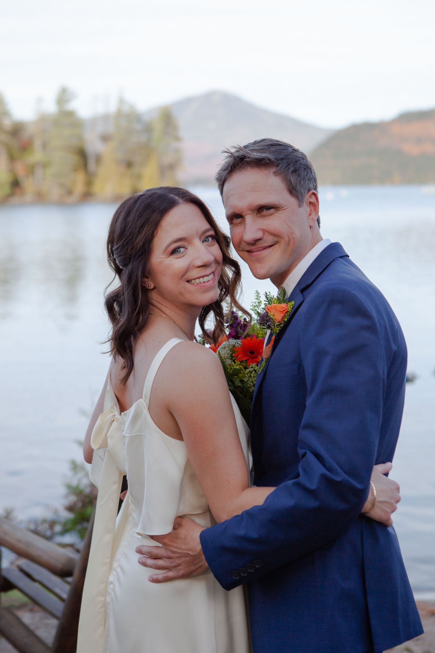 A smiling bride and groom embrace near a lakeside dock with mountain scenery in the background.