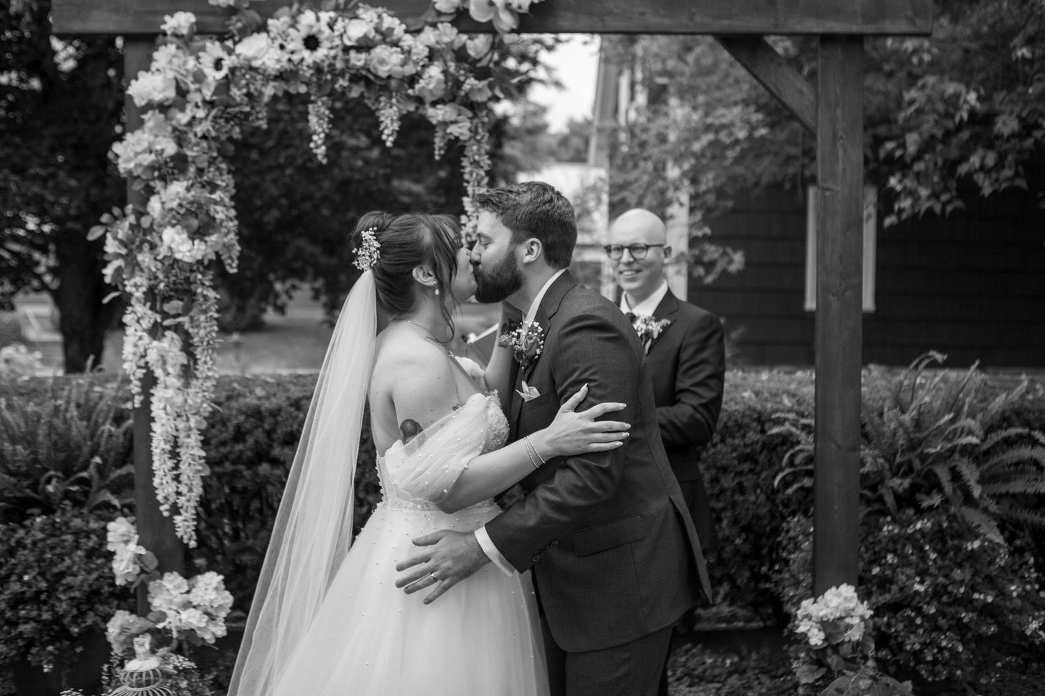 A wedding ceremony under a floral-decorated wooden arch in a garden setting.