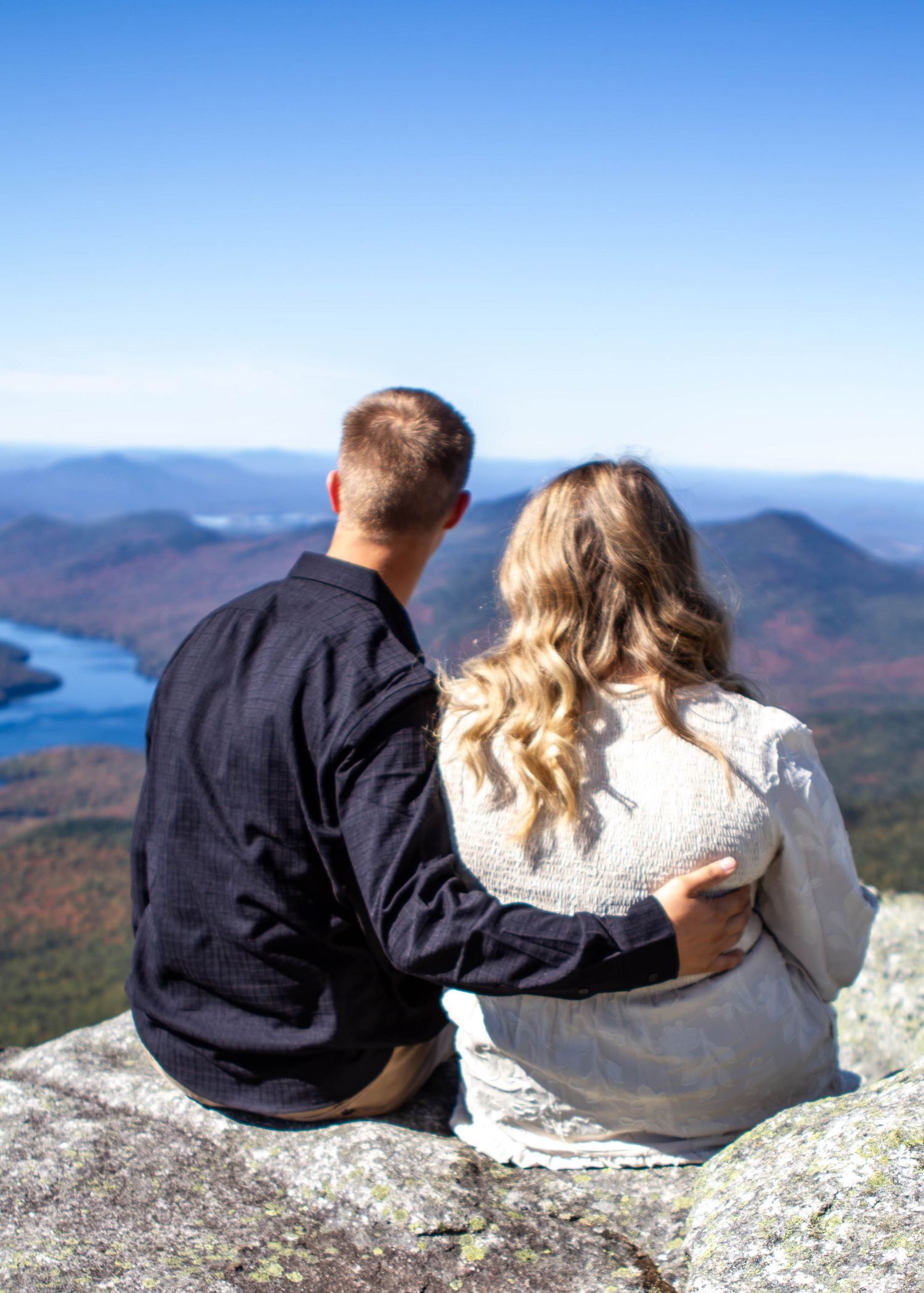 Two people sit together on a mountain overlook with autumn foliage and a lake in the distance.