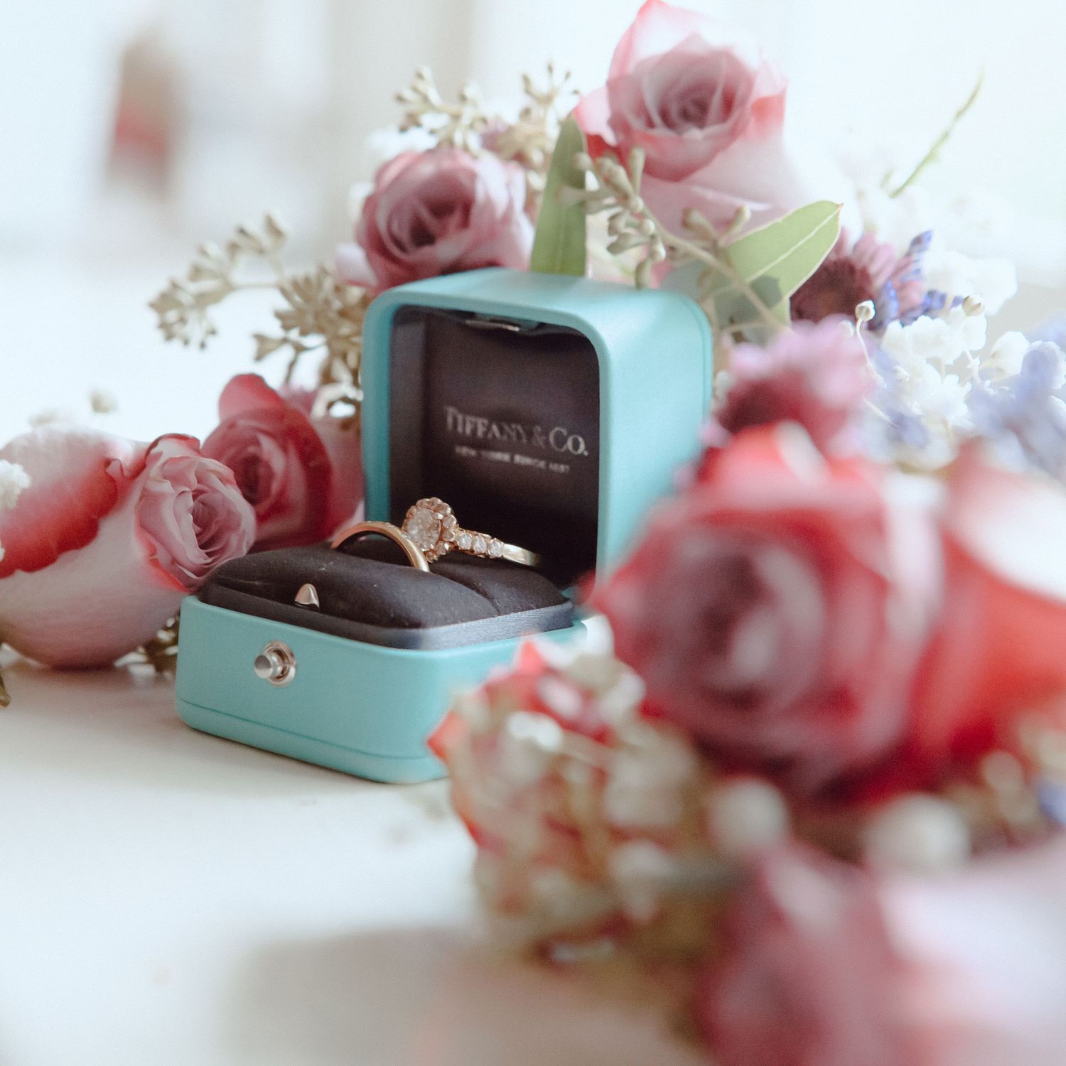 Tiffany engagement ring in turquoise box surrounded by pink roses and delicate flowers.