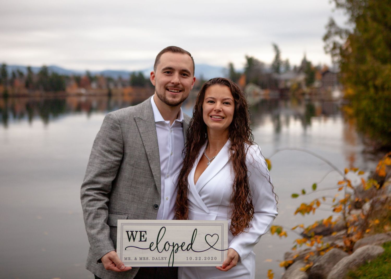 Couple holds 'We Eloped' sign during lakeside photoshoot with mountain backdrop.