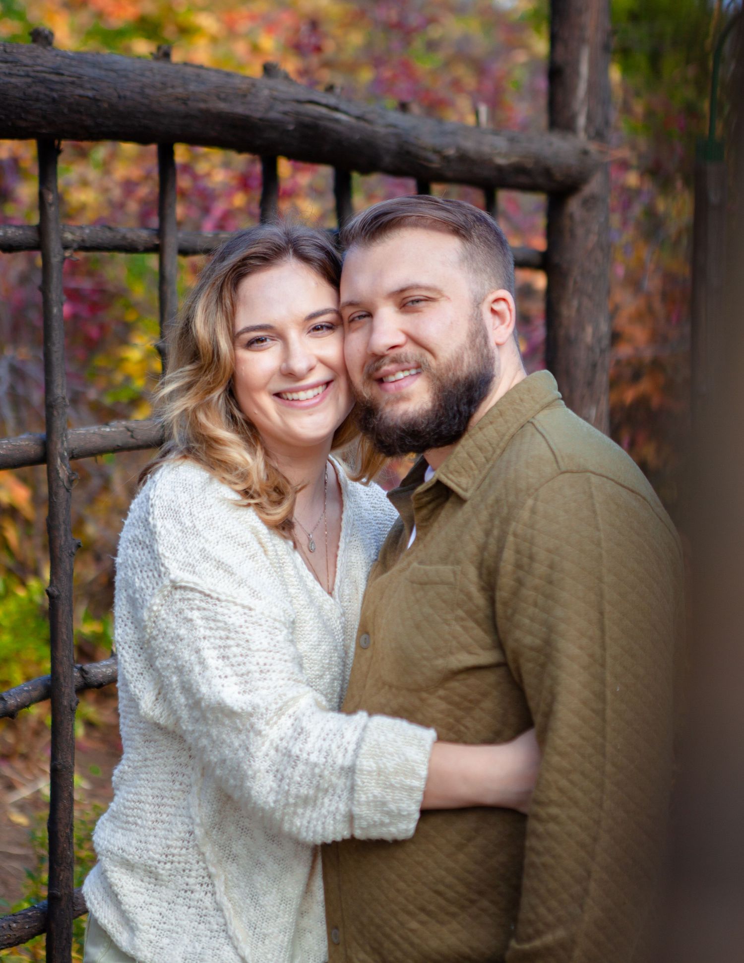 Cozy autumn engagement photos with couple in white sweater and olive shirt against fall foliage.
