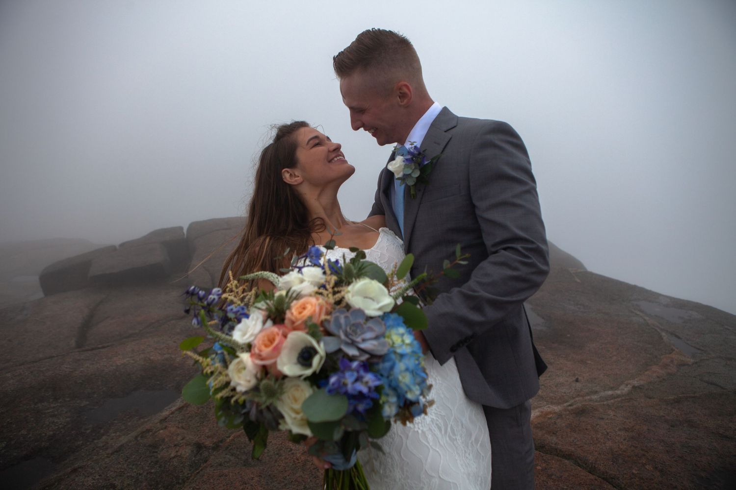 Newlyweds with colorful bouquet embrace in misty mountain setting.
