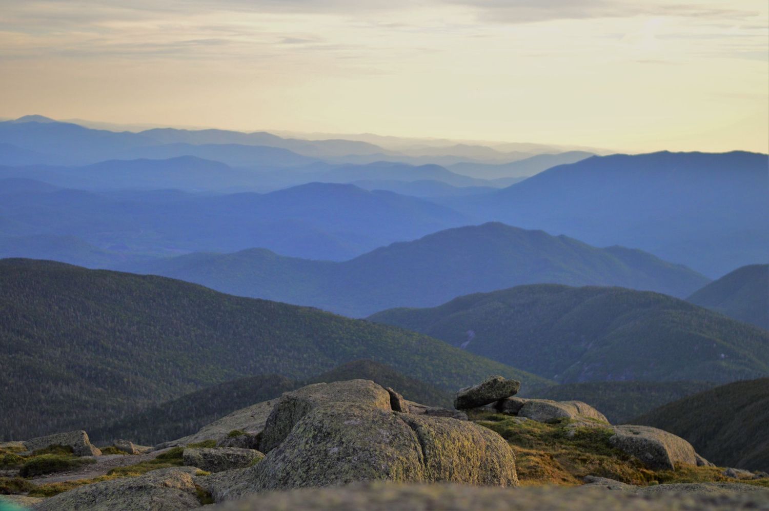 Panoramic view of layered blue mountain ridges at dusk from a rocky summit in a vast wilderness landscape.