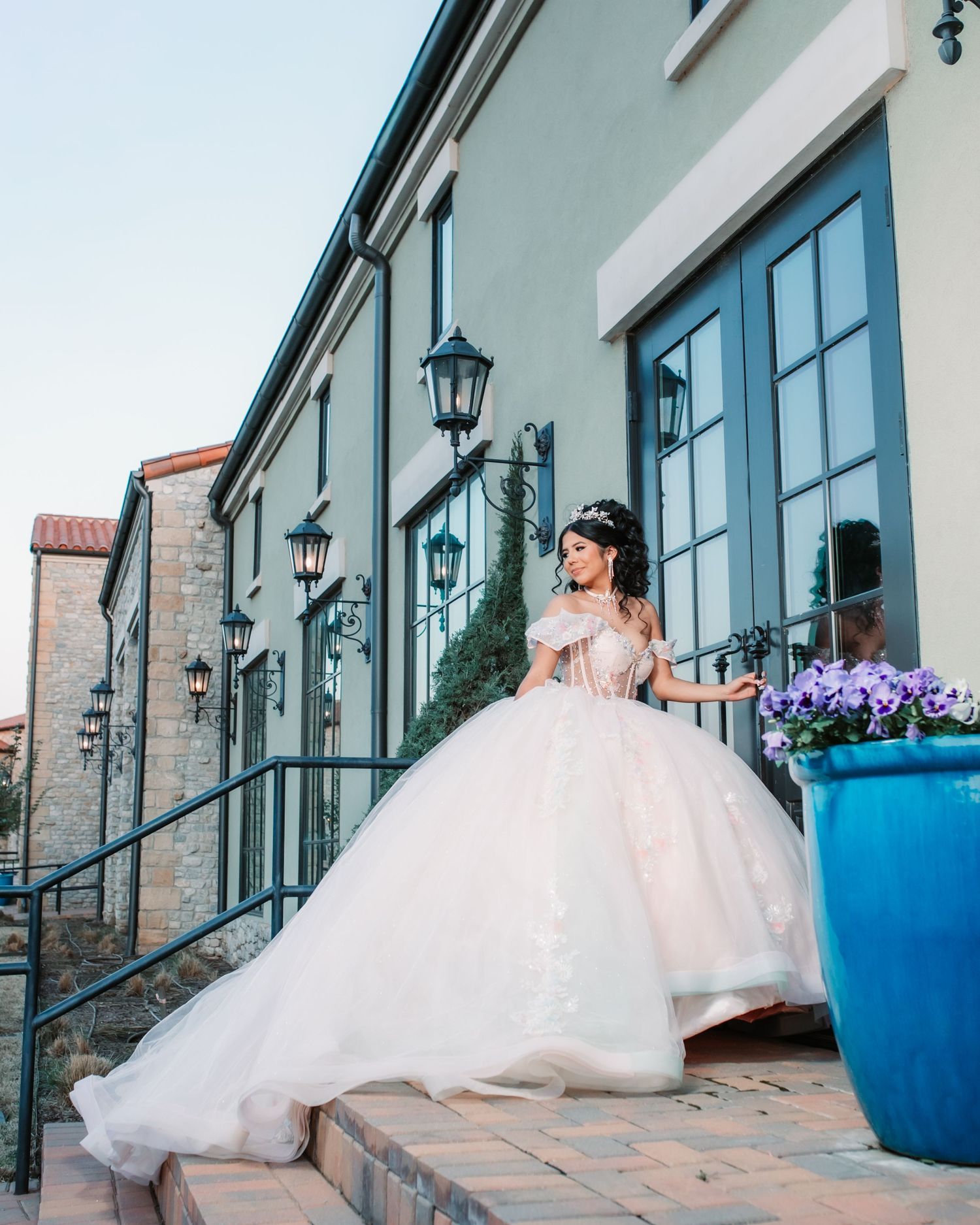 A flowing white ballgown sweeps across stone pathways beside blue planters.