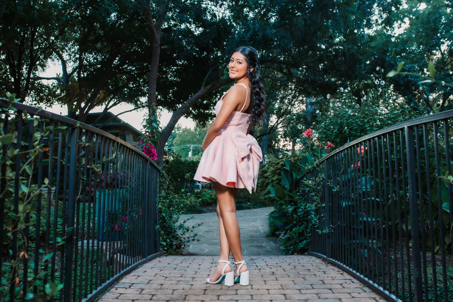 A person in a pink dress walks along a stone pathway surrounded by greenery and iron railings.