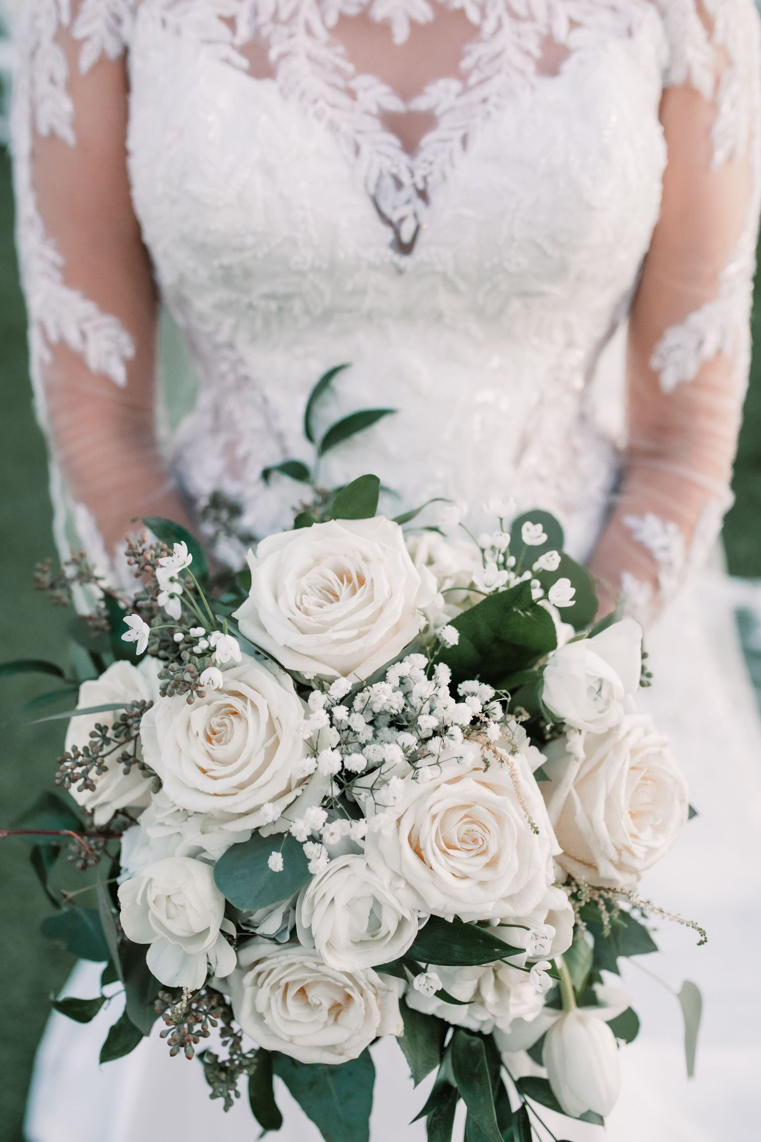 Close-up view of elegant white rose wedding bouquet with baby's breath and eucalyptus greenery against lace dress details.