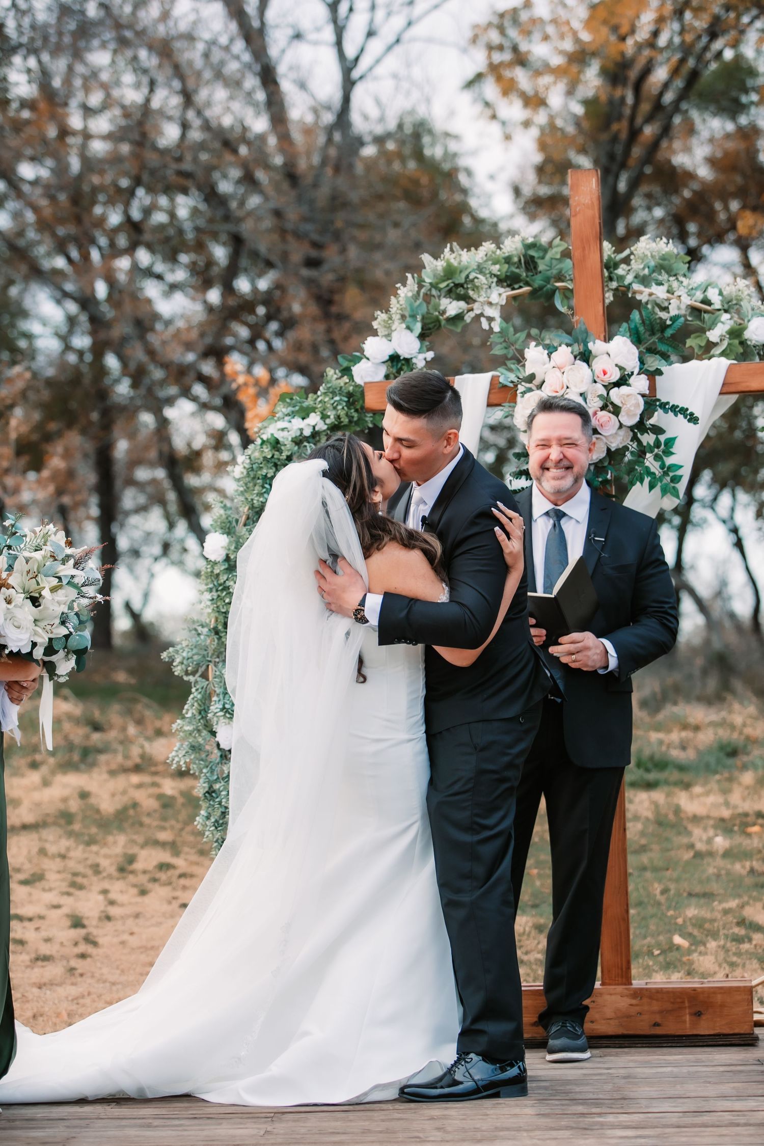 Romantic moment during outdoor ceremony beneath wooden cross decorated with white flowers.