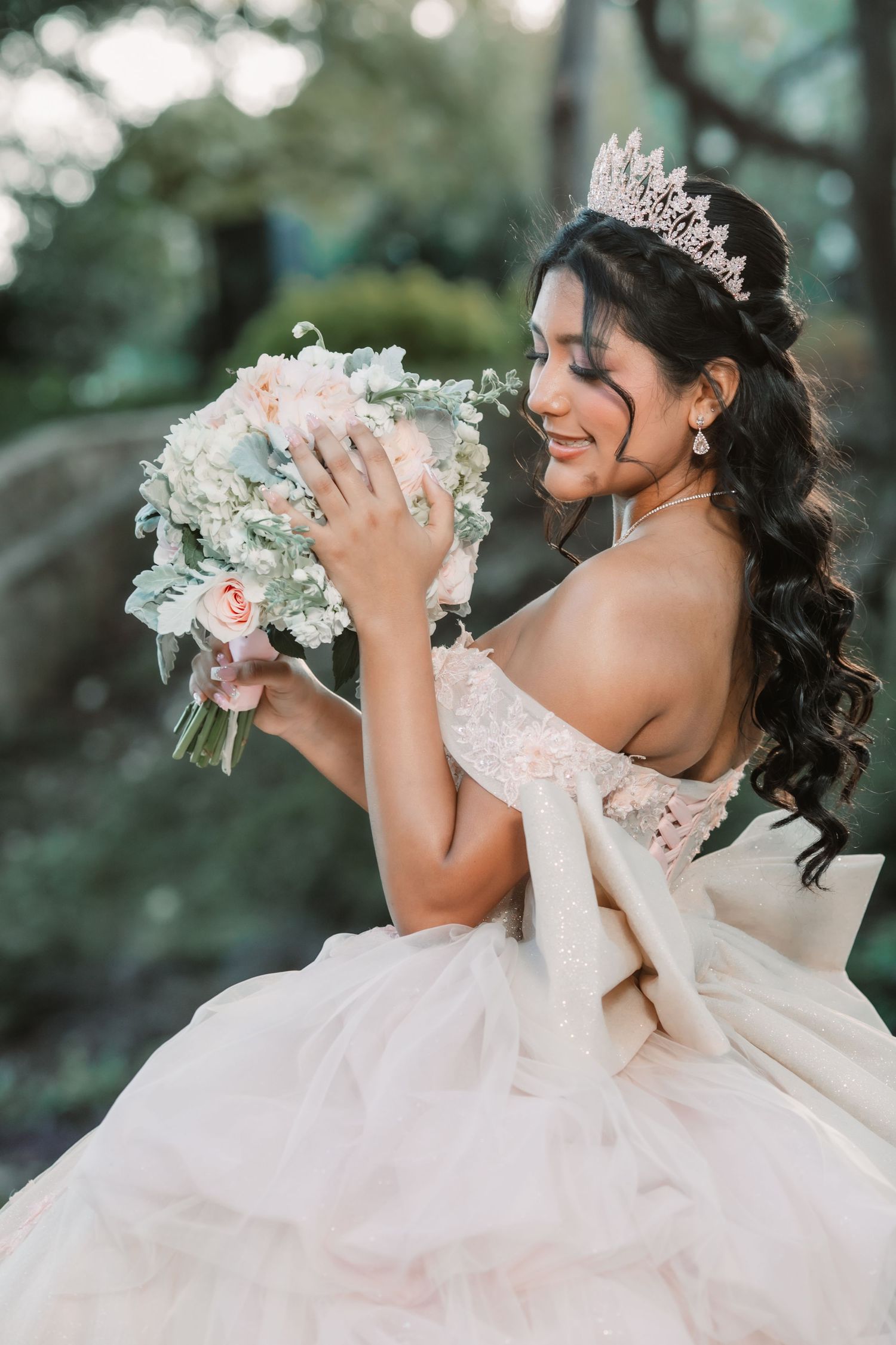 A Quinceañera in a flowing white gown and tiara holds her bouquet against a dreamy bokeh forest background.