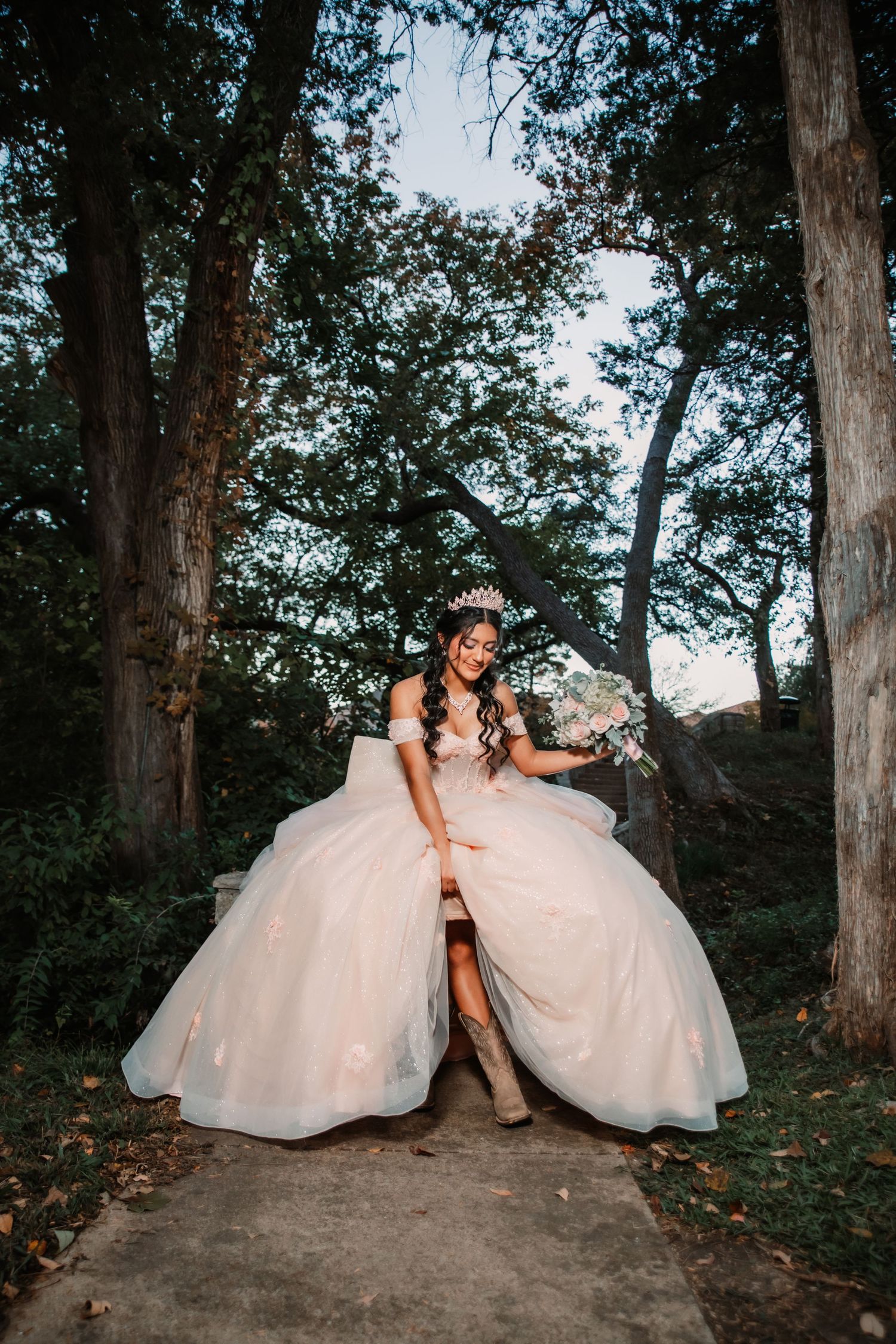 A Quinceañera in a voluminous dress poses gracefully on stone steps in a forest setting.