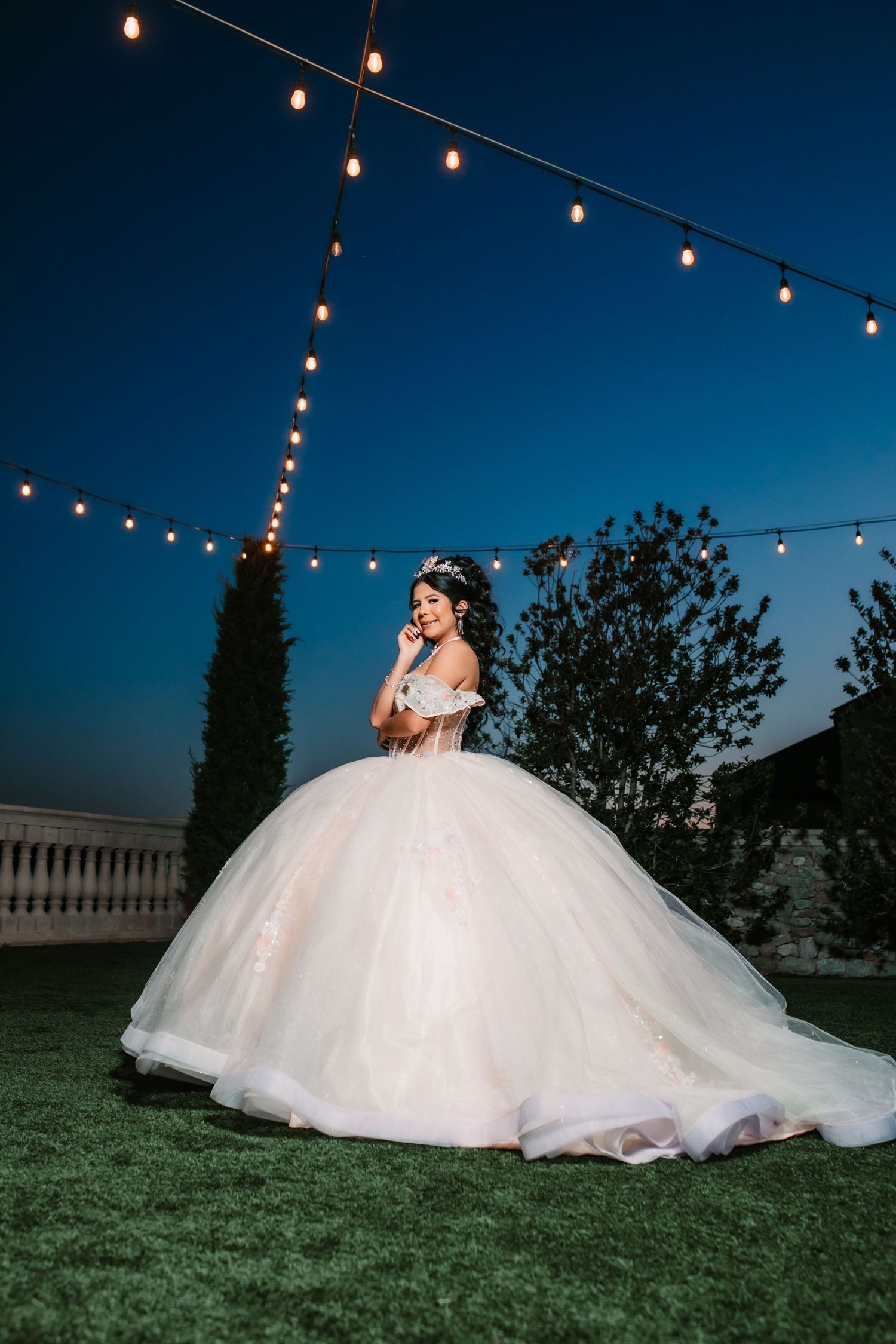 Ethereal Quinceañera portraits at dusk with string lights and flowing white ballgown on grassy hill.