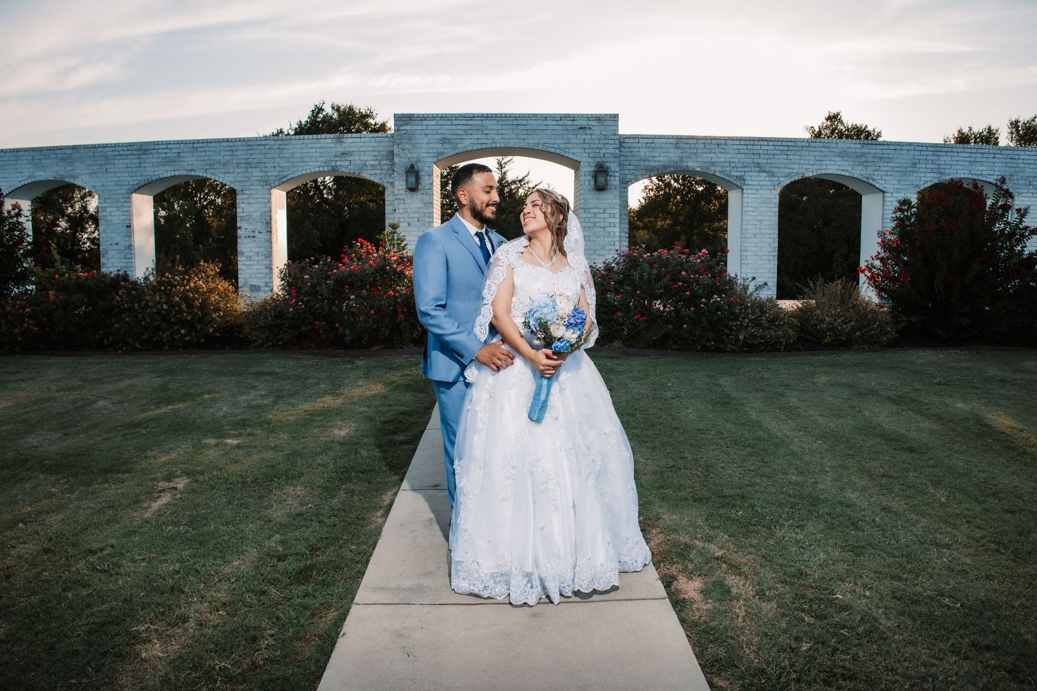 Wedding couple poses in front of a white architectural structure on a grassy lawn at sunset.