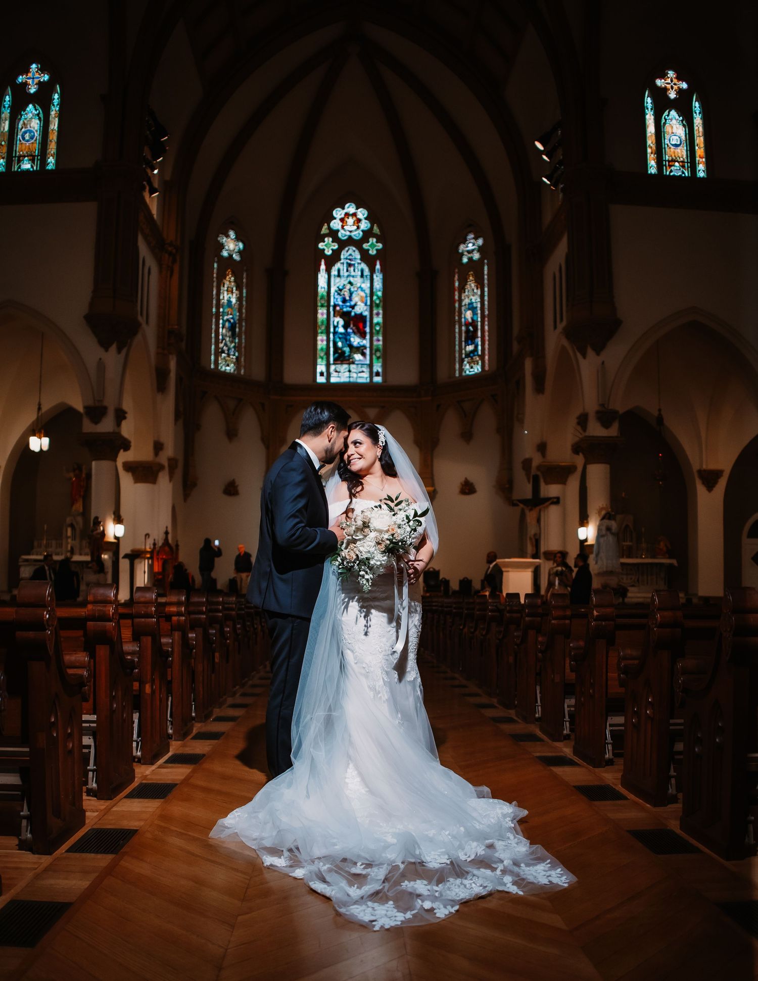 A romantic wedding couple embraces in the center aisle of a grand cathedral with stained glass windows and wooden pews.