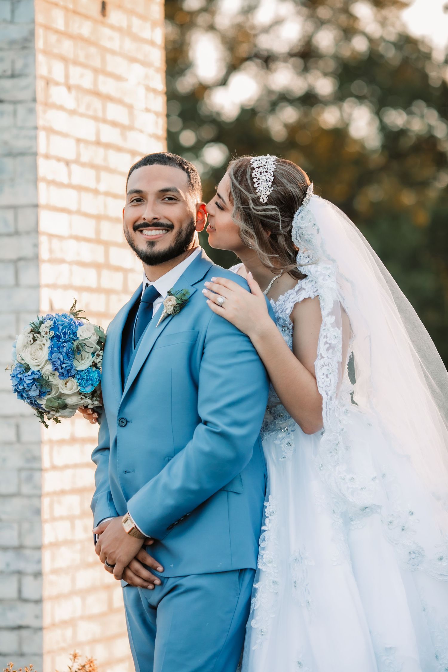 A romantic outdoor wedding photoshoot featuring a light blue suit and white gown against a brick wall backdrop.