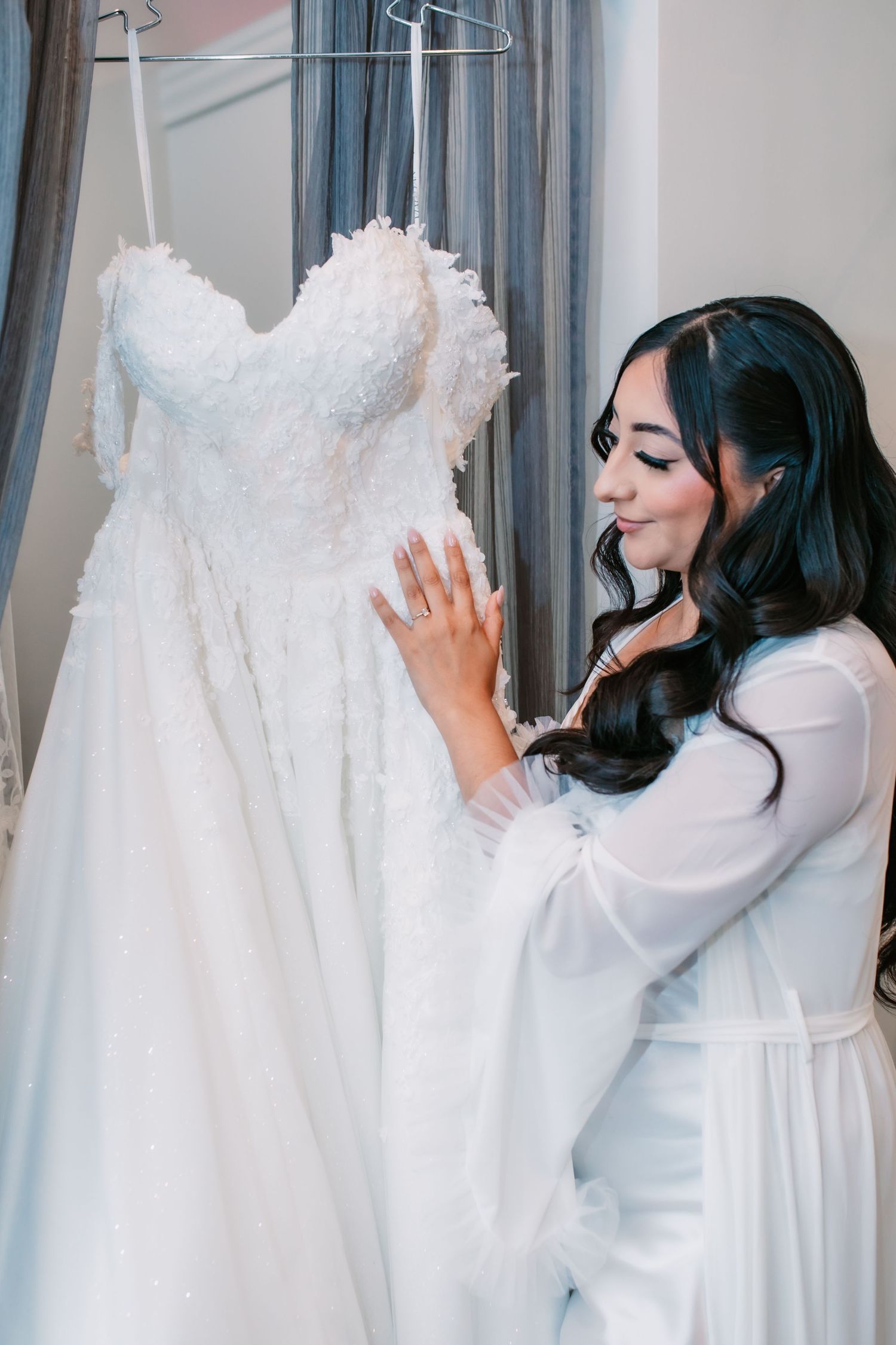 A beautiful white lace wedding dress hangs on display as someone in white admires its intricate details.