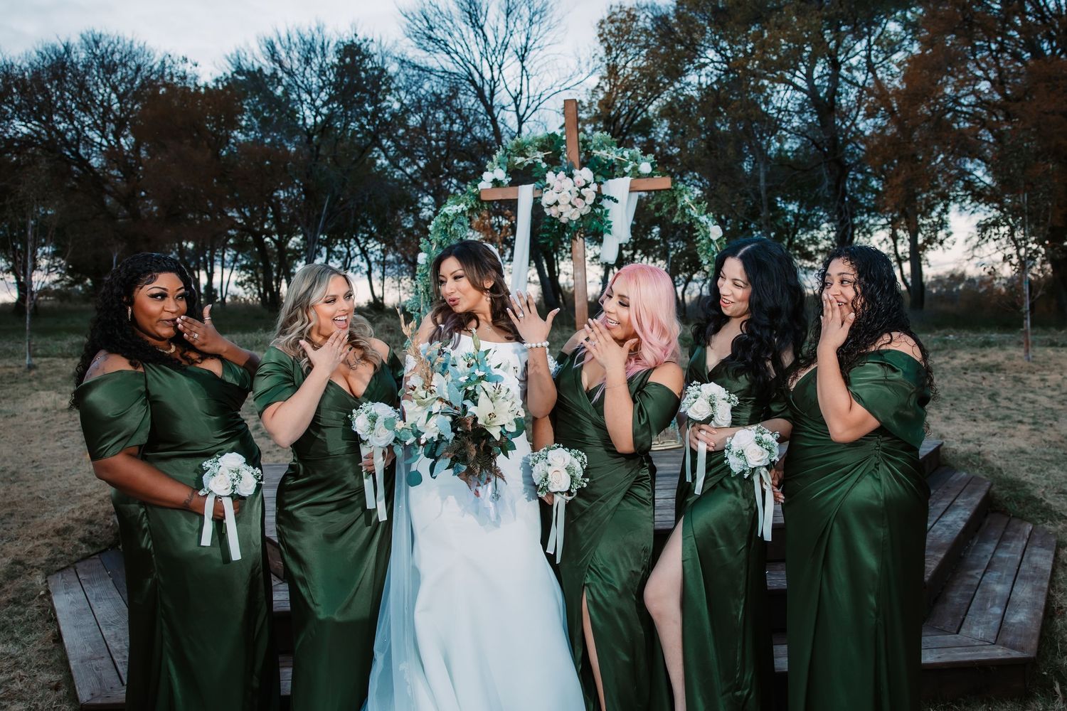 Bridesmaids in emerald green dresses pose playfully with bouquets at outdoor wedding.