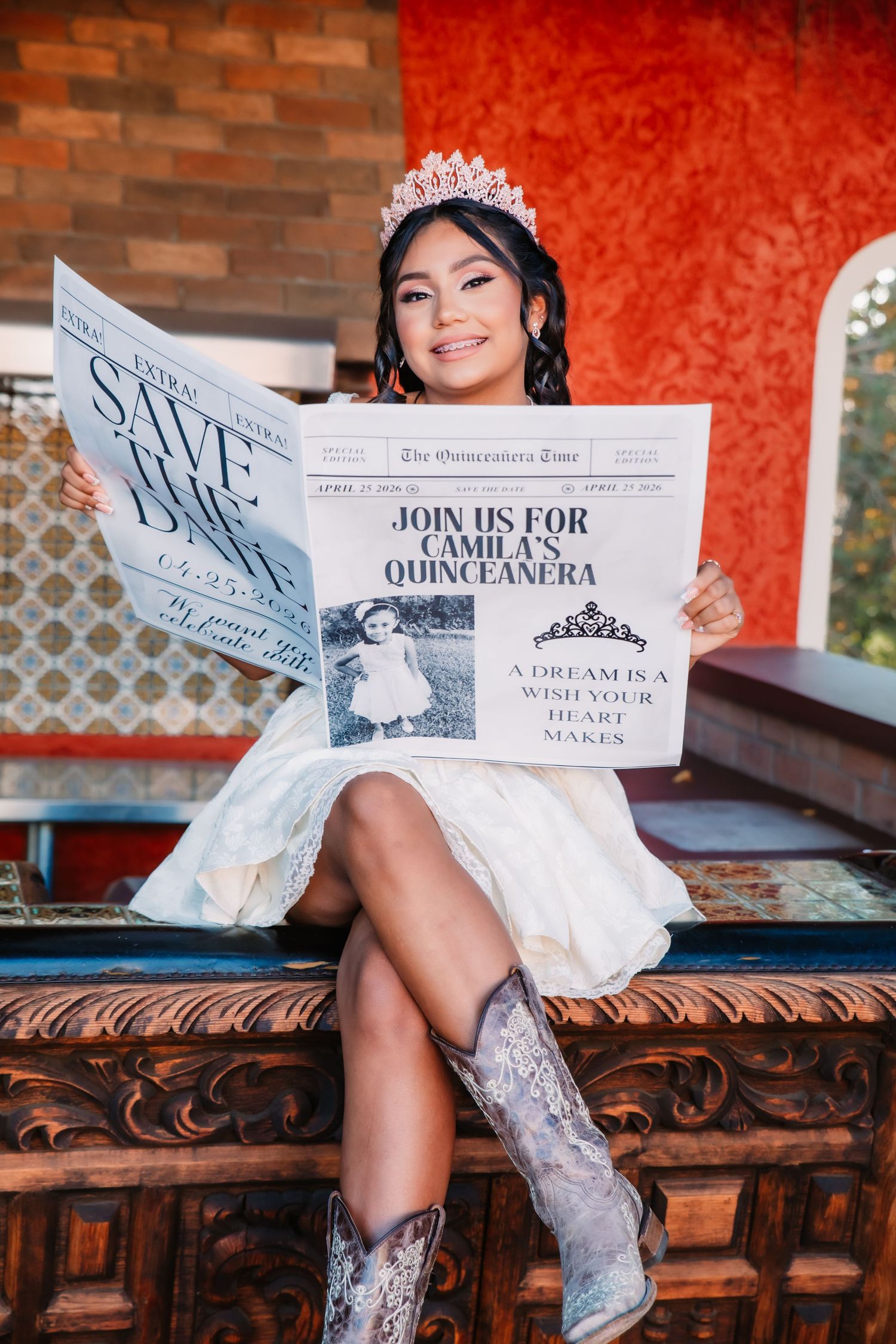 A person in a white dress and cowboy boots poses with a newspaper on an ornate wooden bench.