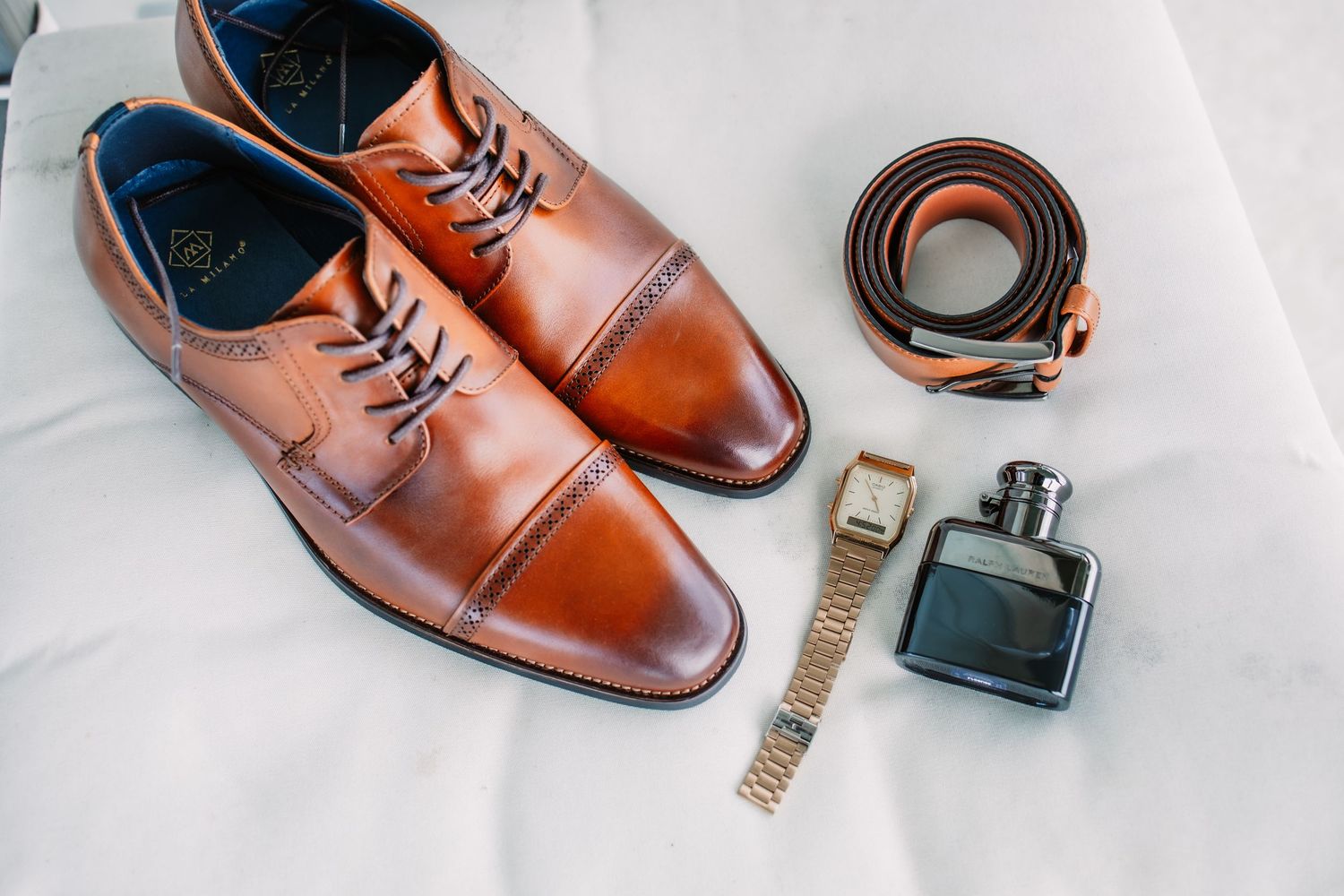 Brown leather dress shoes and accessories laid out on white surface for wedding day.
