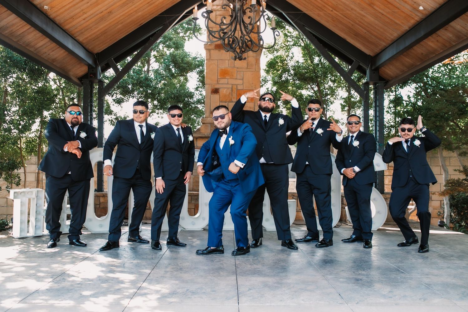 Groomsmen in black suits pose with the groom in blue under a wooden pavilion.