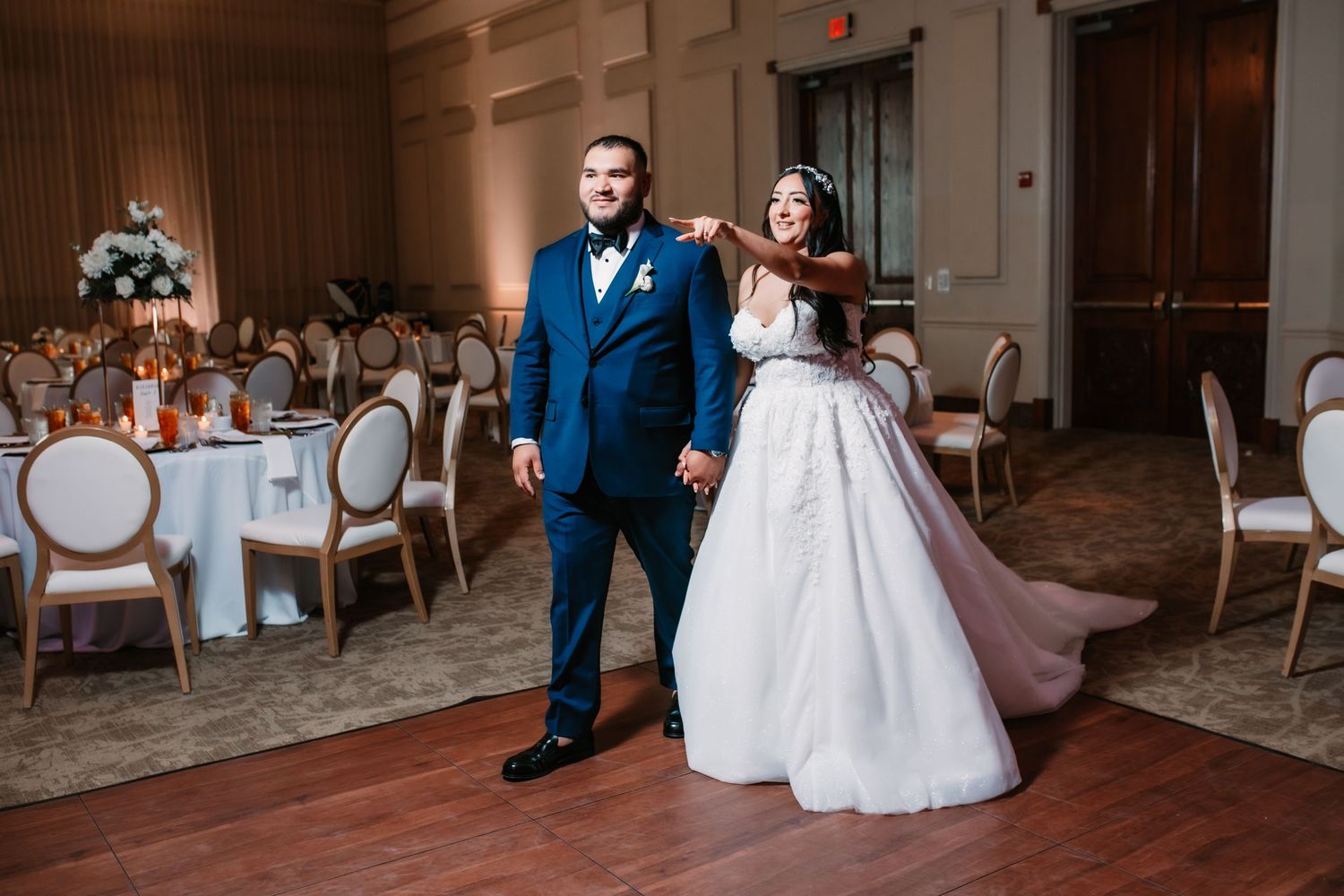 A couple poses dramatically in an elegant reception hall with round tables and classic decor.
