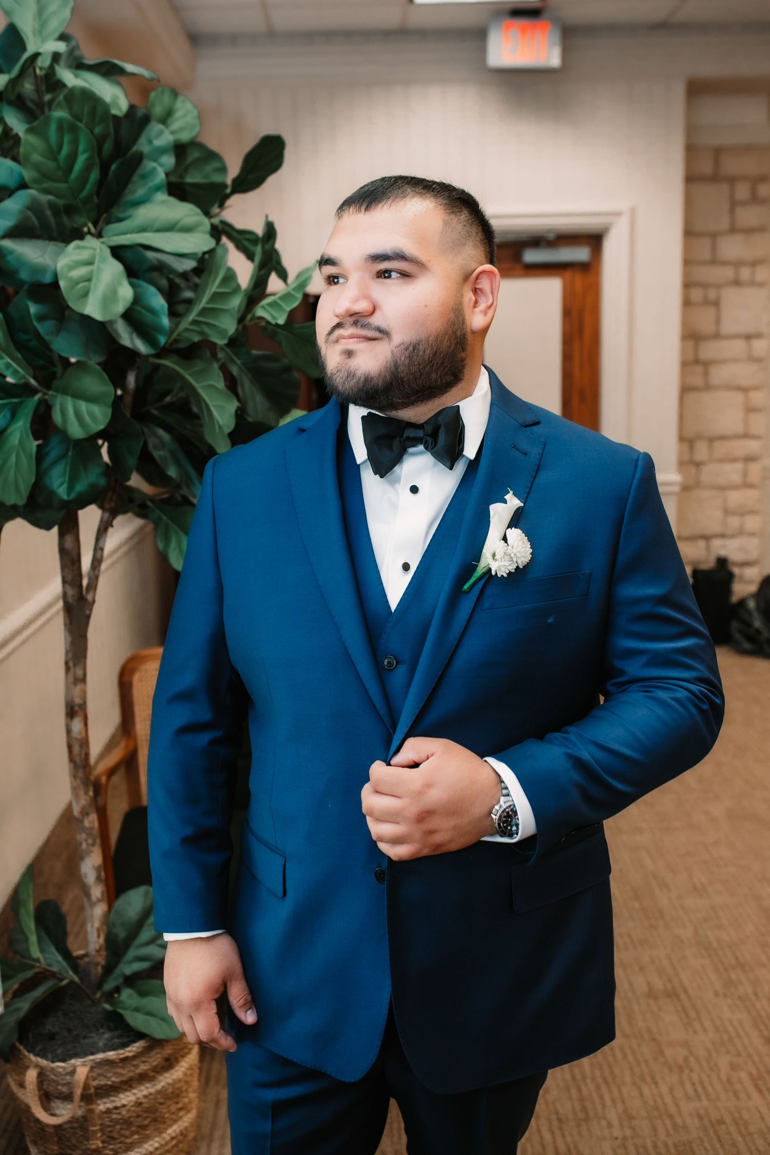 A groom in a classic navy blue tuxedo and bowtie poses elegantly next to a potted plant.