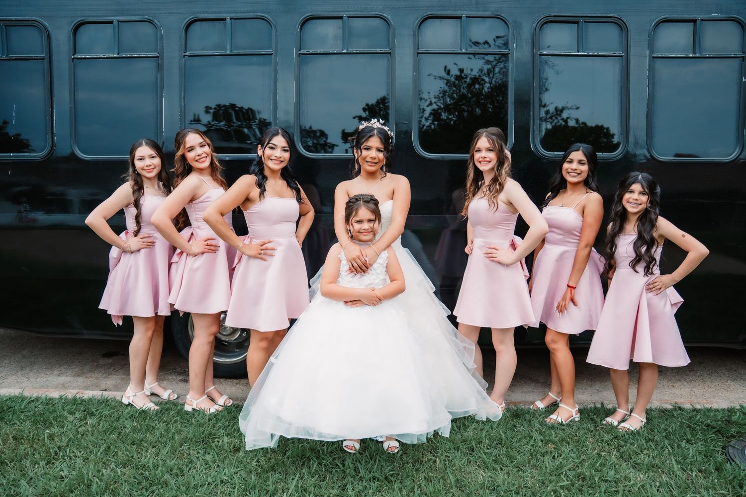 Quince party in pink dresses posed casually in front of vintage bus.