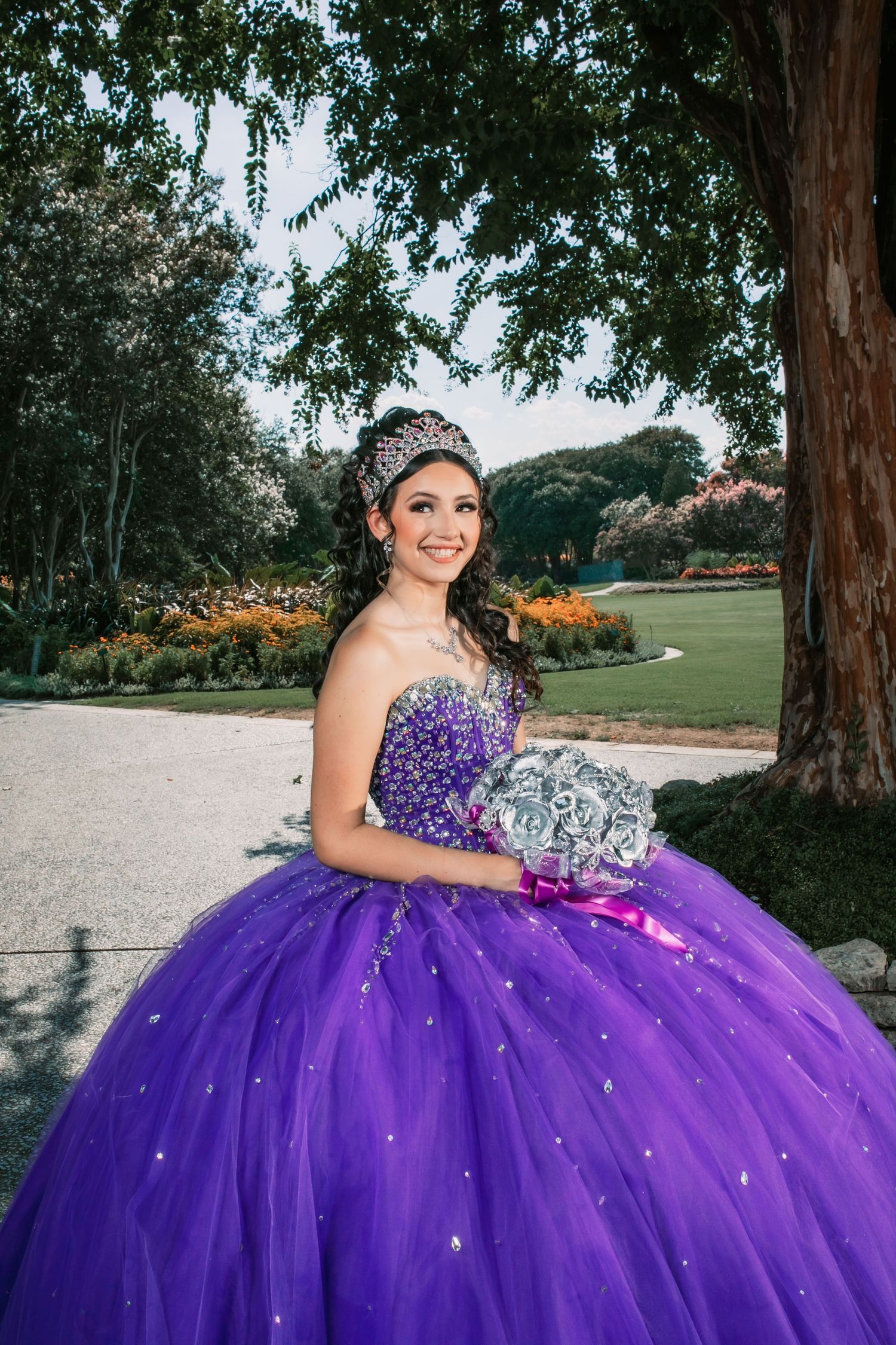 A quinceañera celebration featuring a stunning purple ball gown with sparkles and a crystal tiara in an outdoor garden setting.