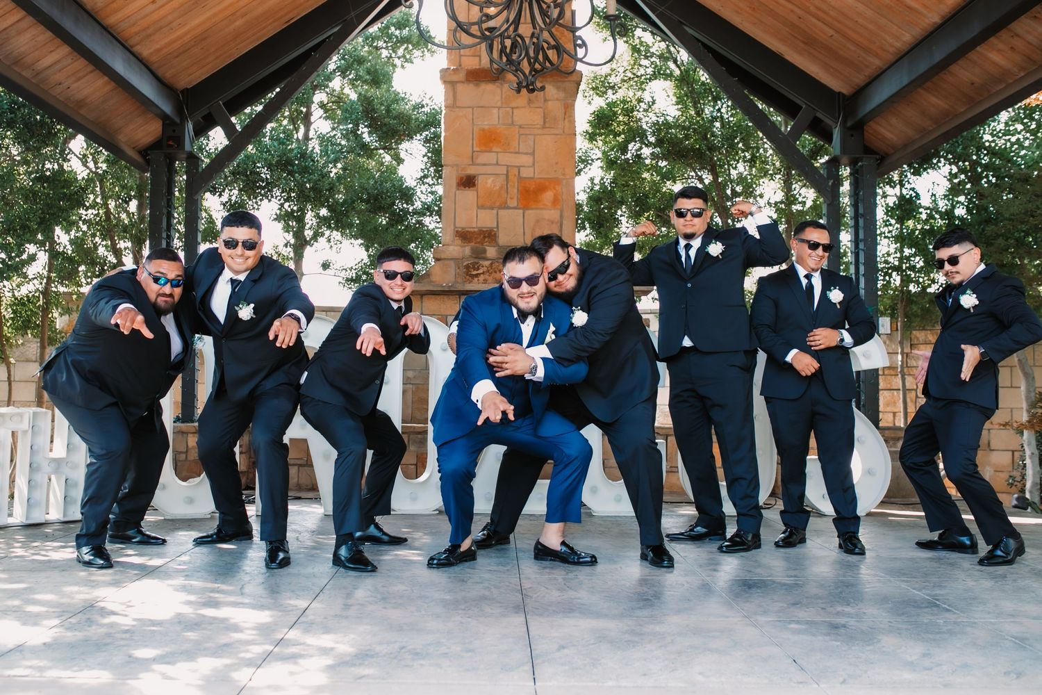 Groomsmen strike fun poses with the groom under a covered outdoor venue.