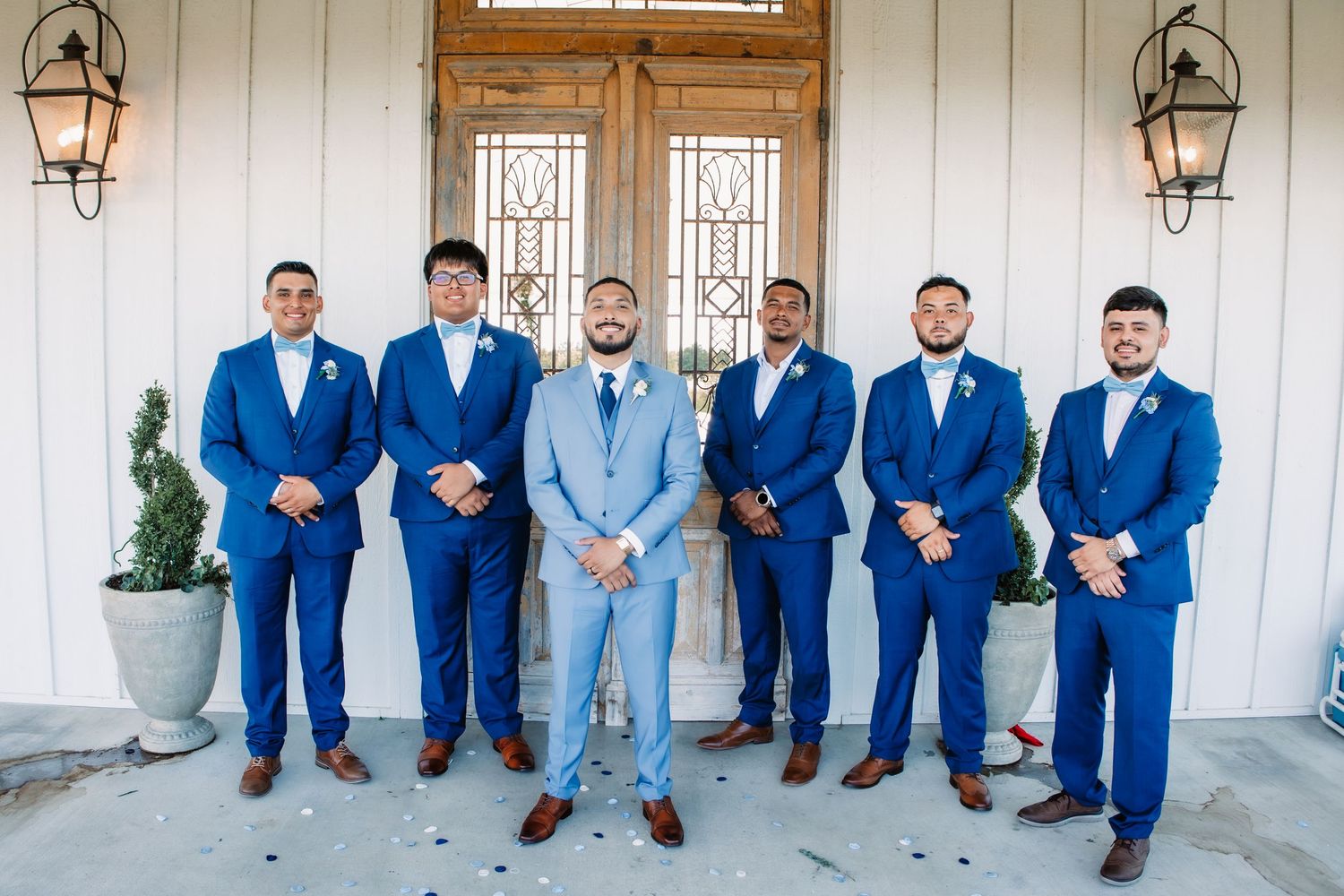 Wedding party in coordinating blue suits posed against white building exterior.