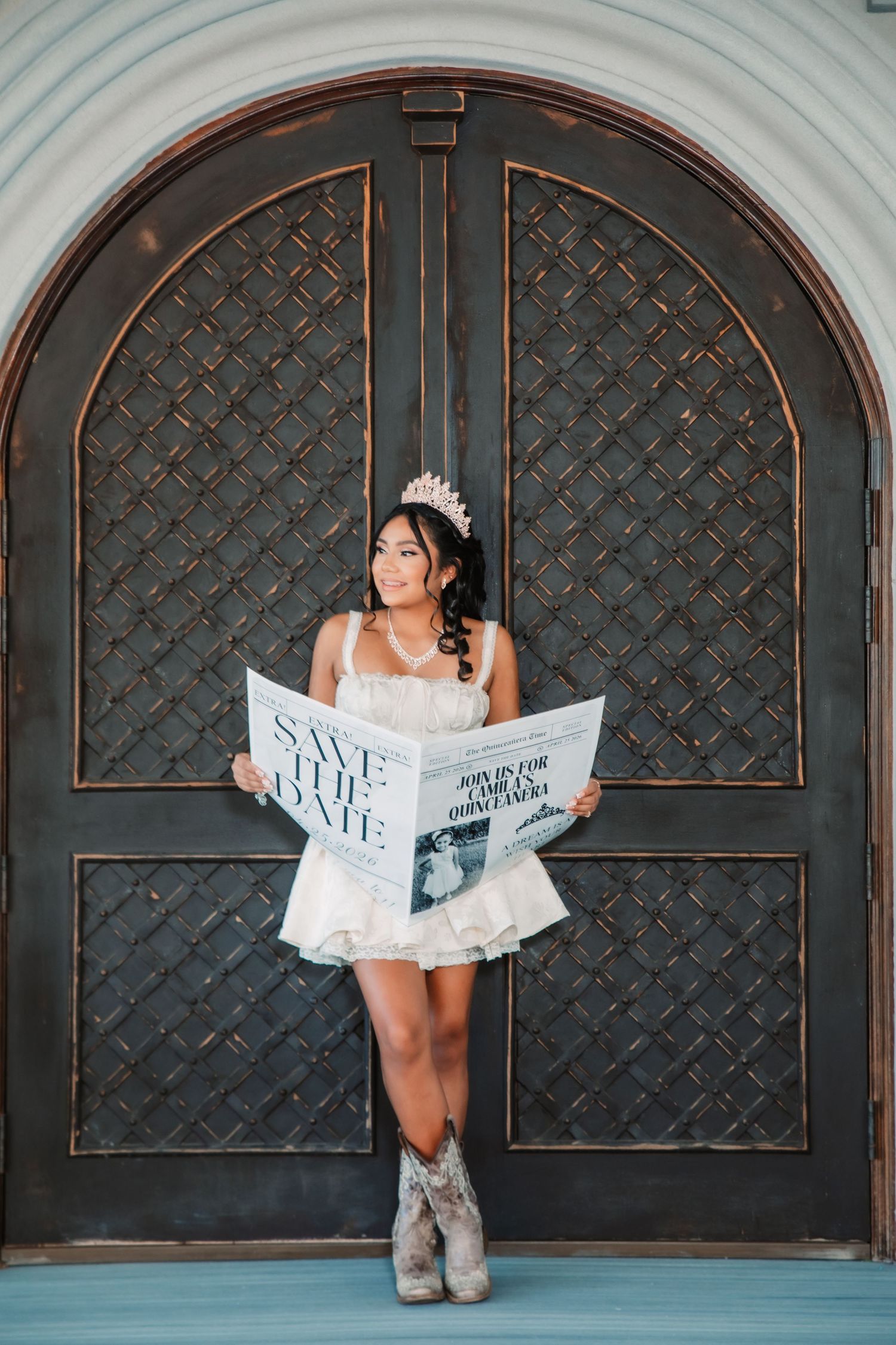 A person in a white dress and boots poses against dark patterned doors while holding a newspaper.