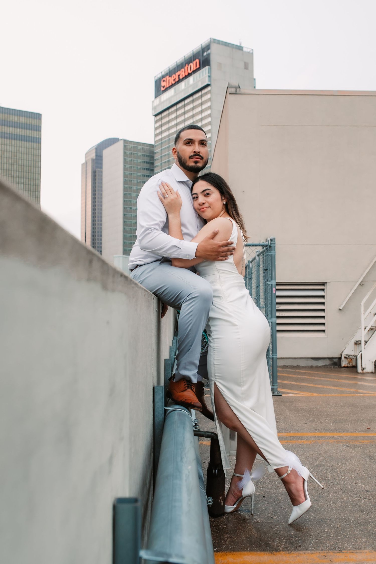 Couple in white and grey attire embracing on urban rooftop with city views.