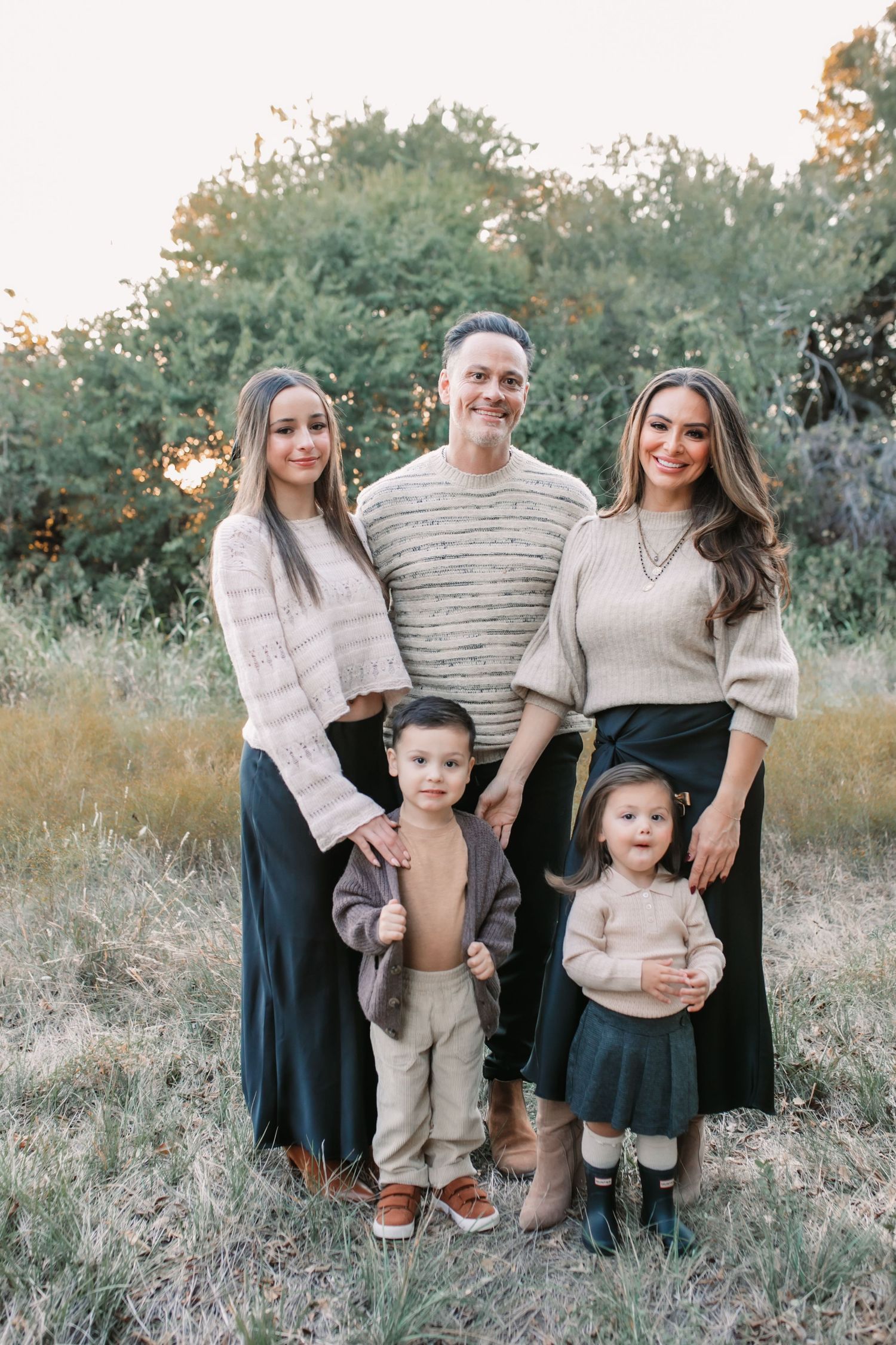 Family of five poses outdoors in coordinated neutral outfits against natural foliage background at sunset.