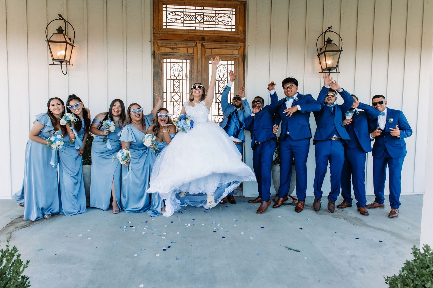 Large wedding party in blue and white attire posed together against white building exterior.