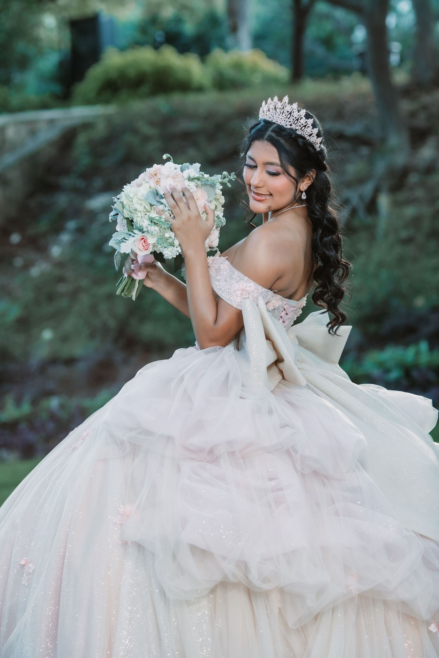 A bride in an elegant off-shoulder ball gown and tiara holds a pink and white bouquet in a dreamy forest setting.