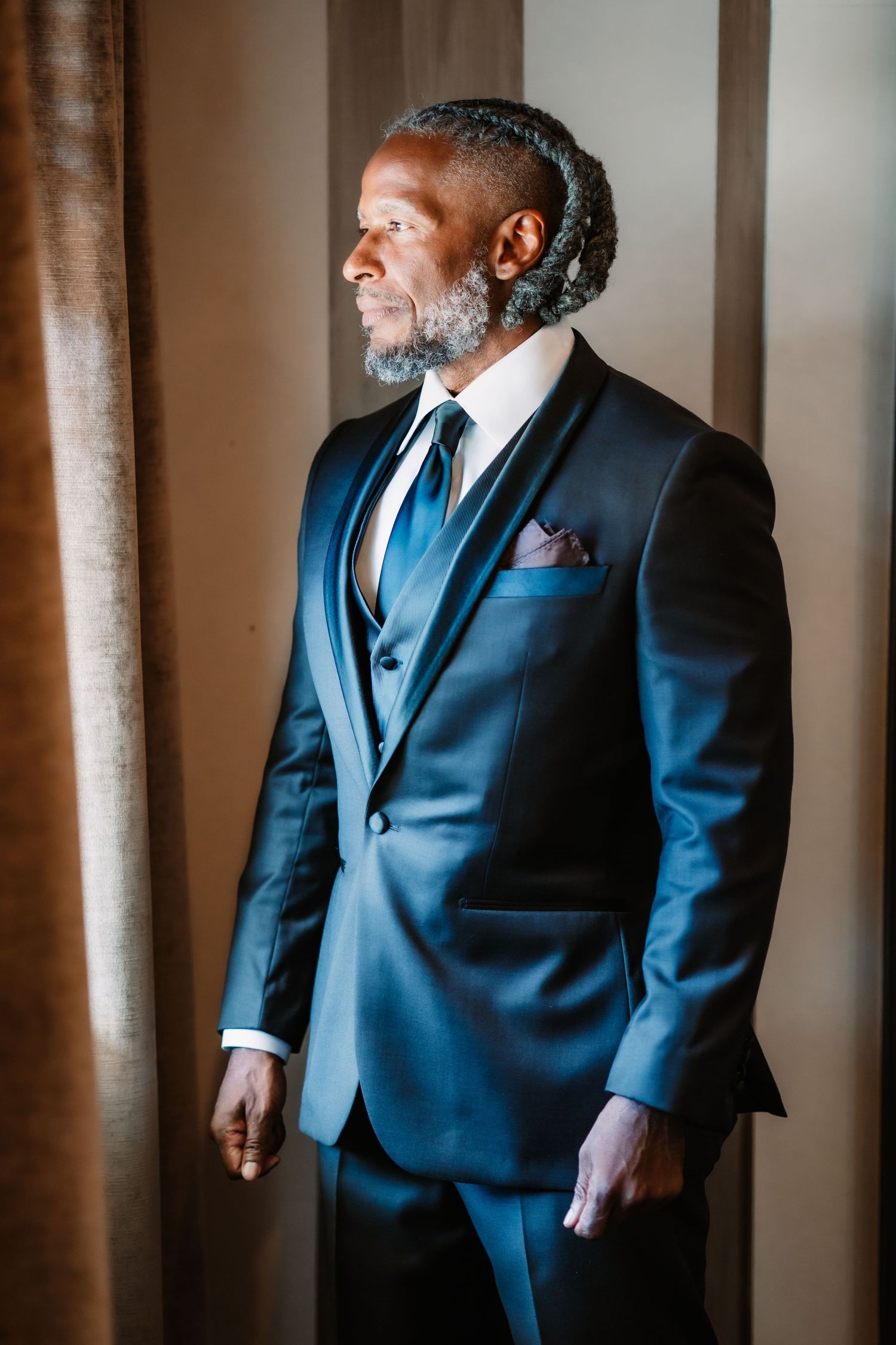 A groom in a navy blue suit and vest stands in dramatic window lighting.