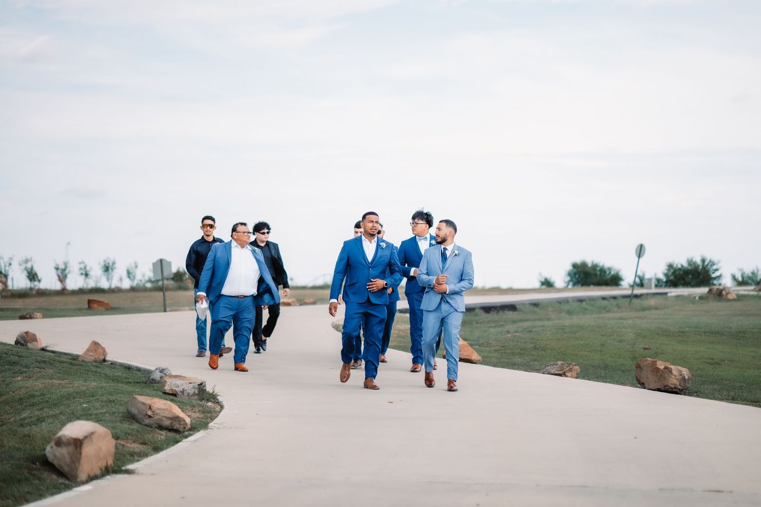 Wedding party in blue suits walking together on a paved outdoor path.