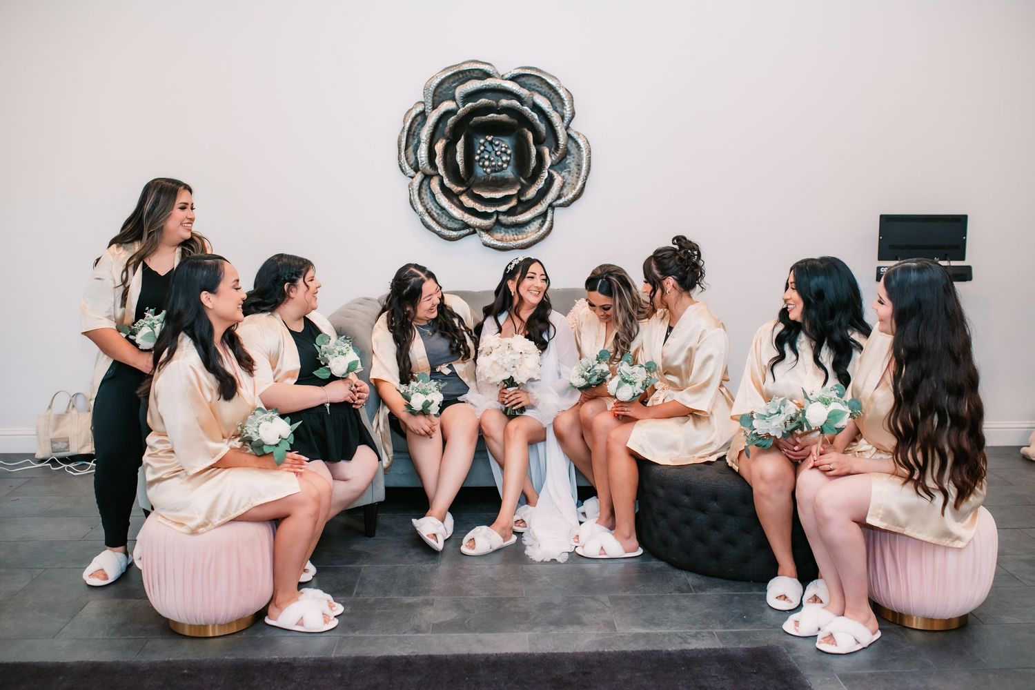 Bridesmaids in matching white robes gather on plush seating beneath a decorative wall flower.