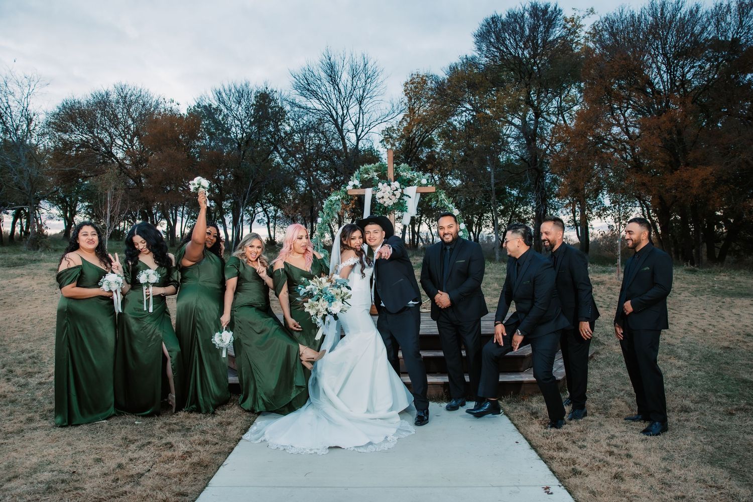 Full wedding party posed on concrete path with bridesmaids in green and groomsmen in black.