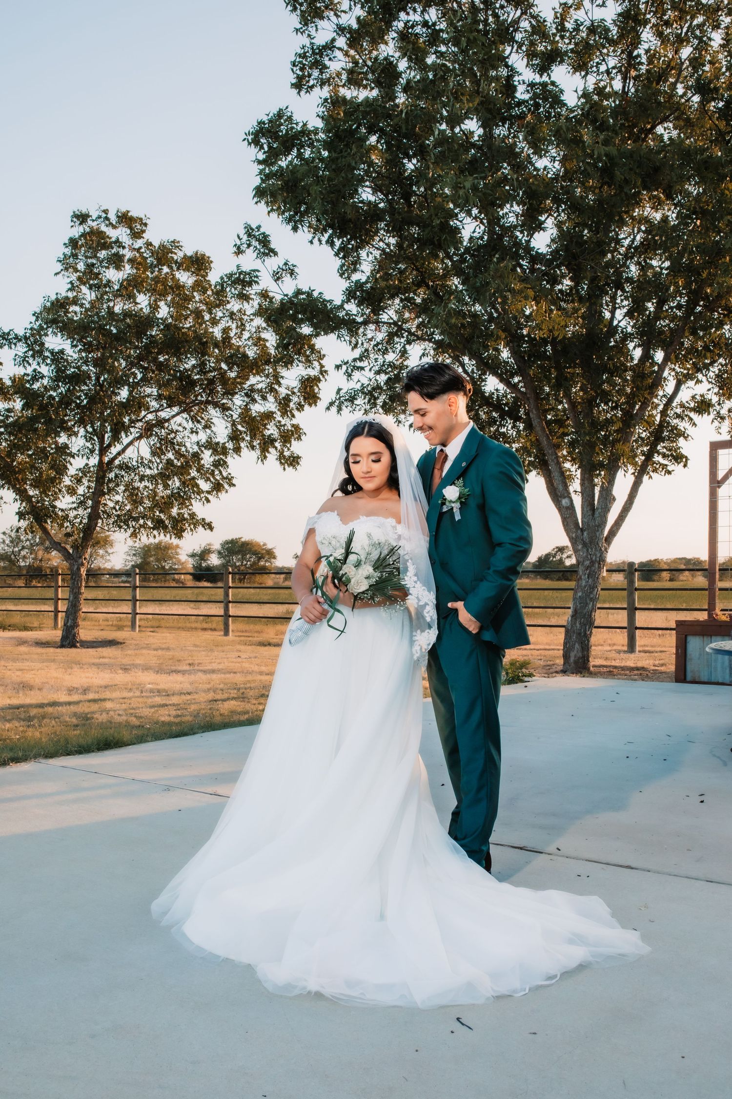 A couple in wedding attire pose together outdoors with trees and a rustic fence in the background at sunset.