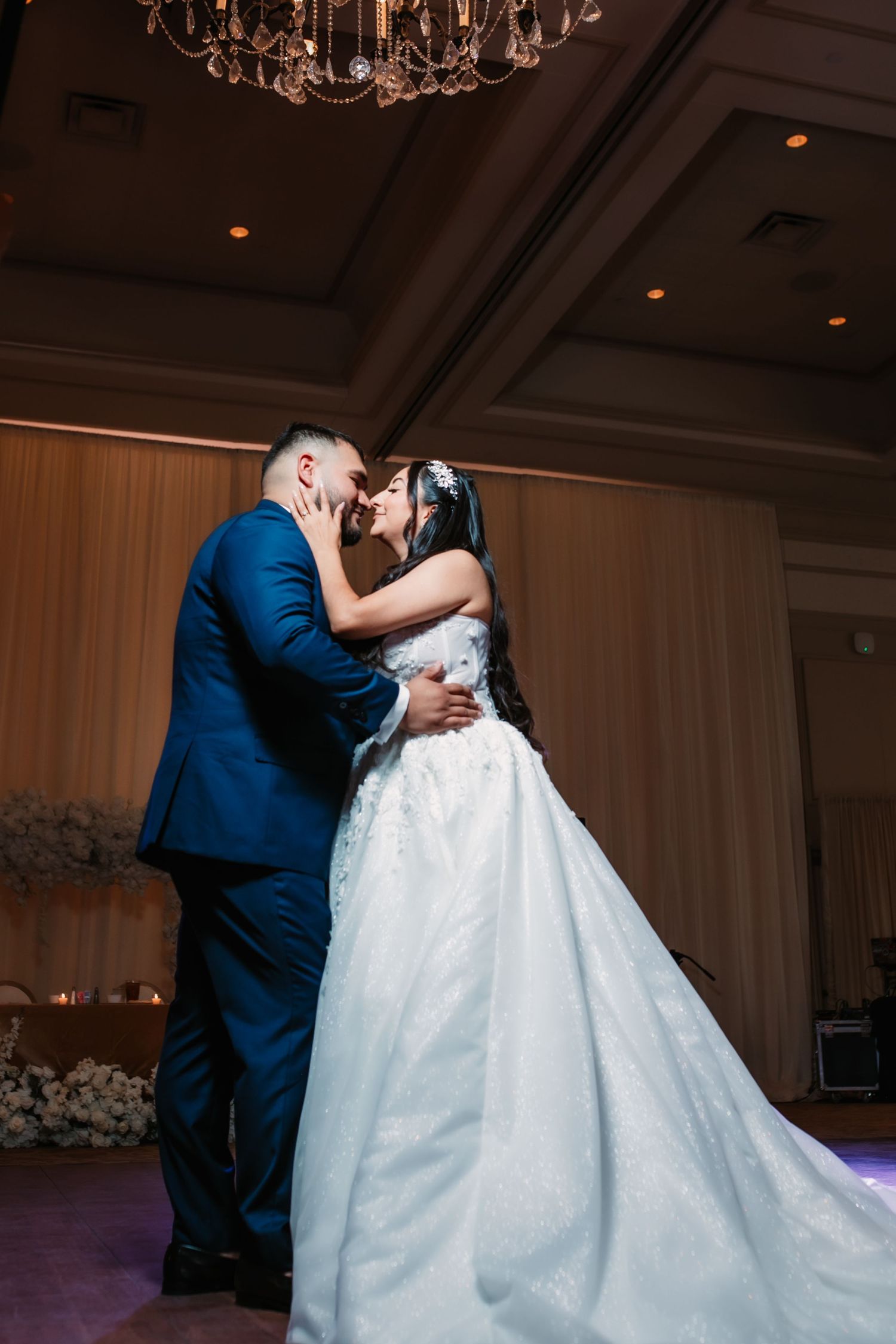 A romantic wedding dance moment in an elegant ballroom with chandeliers overhead.