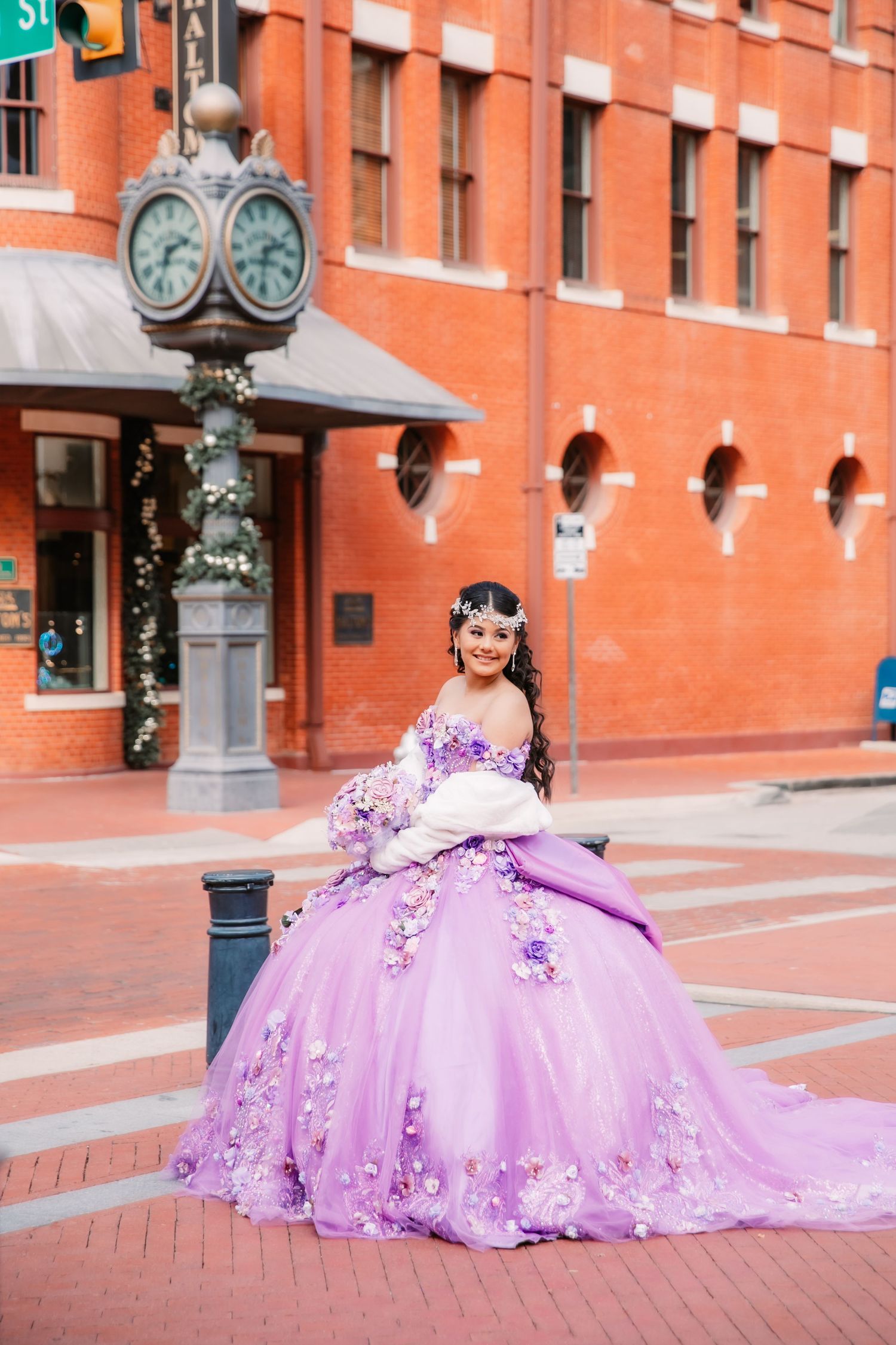 A lavender quinceañera dress flows across red brick pavement near an ornate clock and historic building facade.