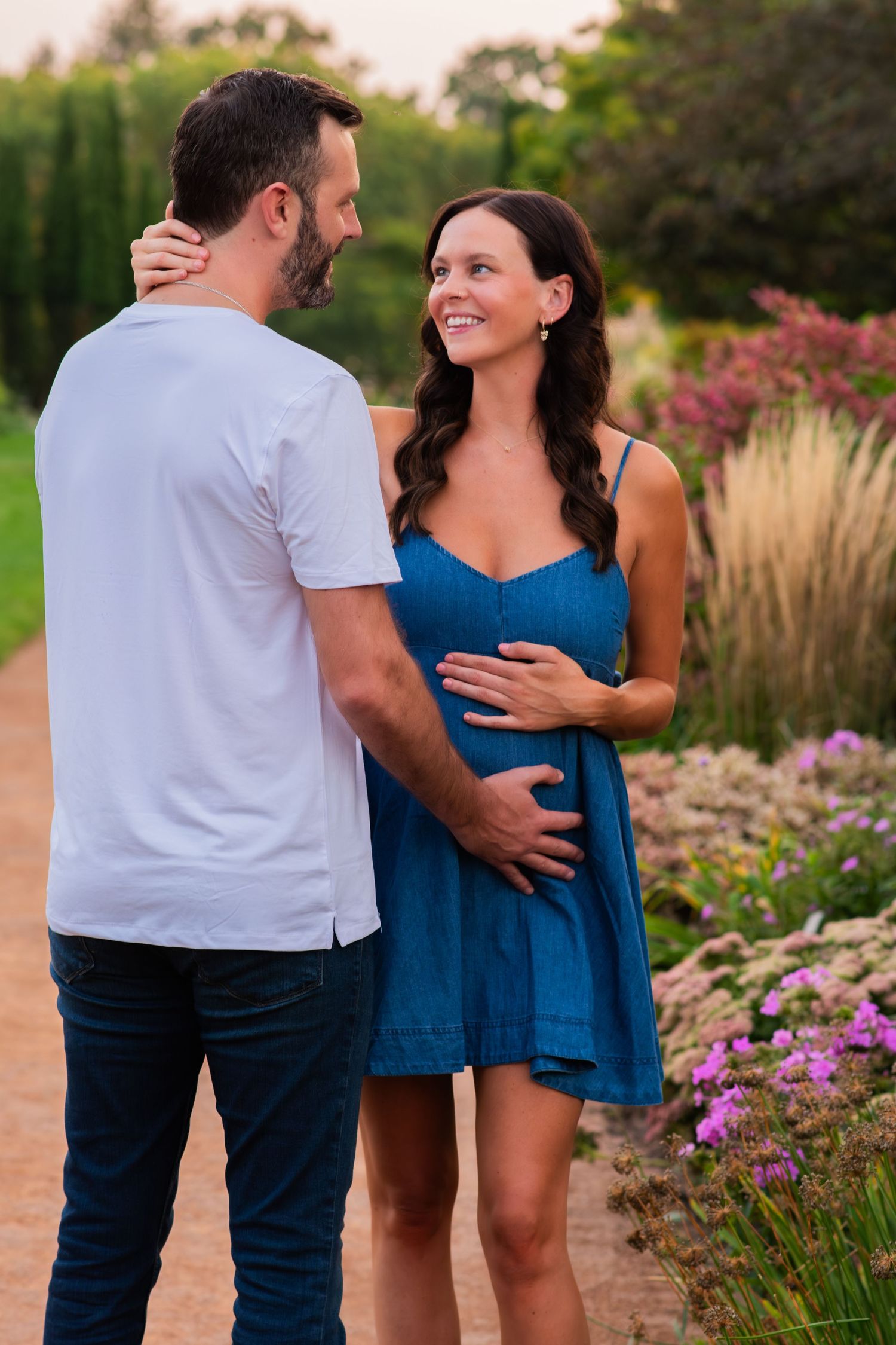 Couple sharing a tender moment in a garden, woman in blue dress smiling while holding her baby bump.